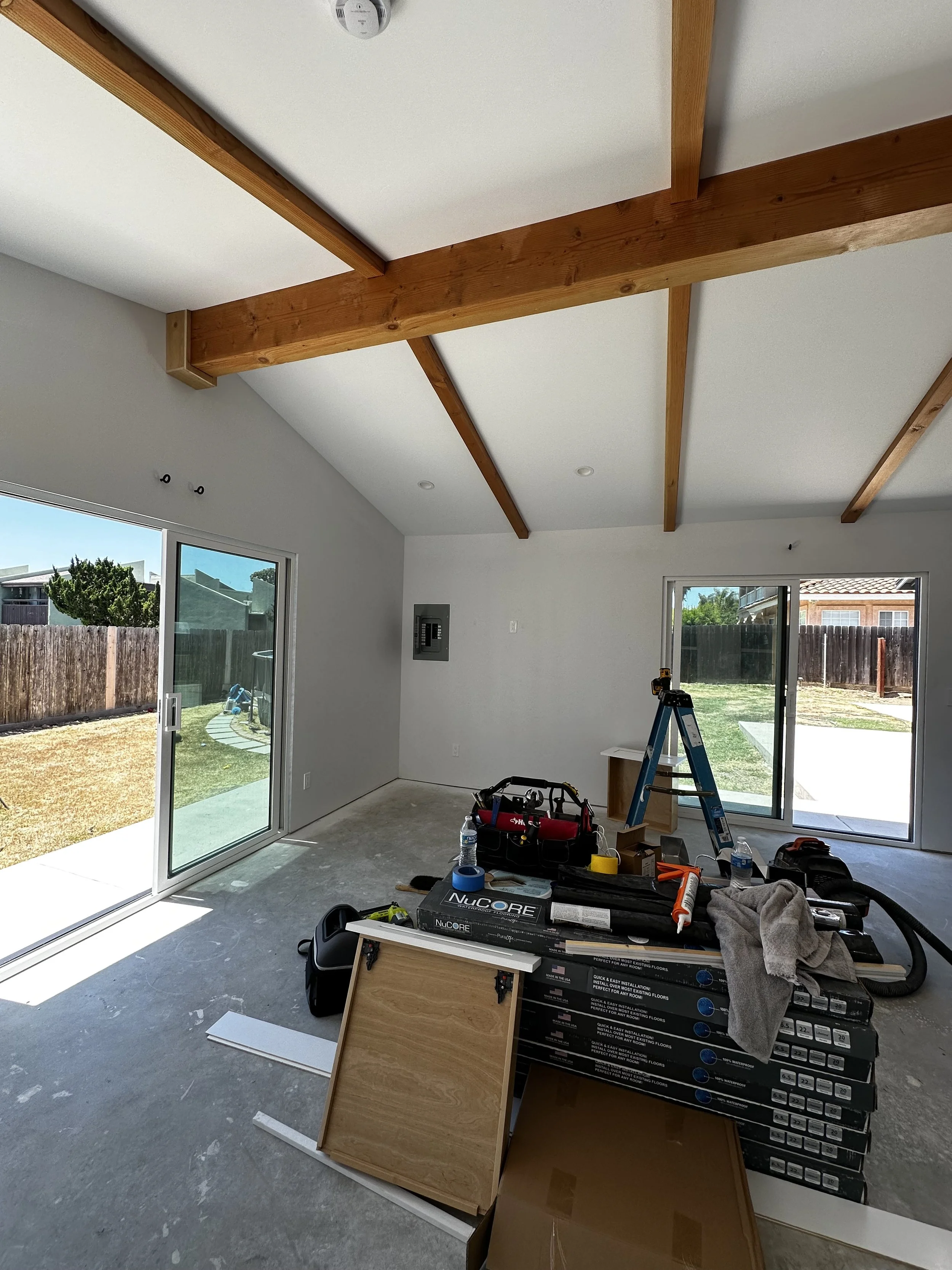 An unfinished room with large sliding glass doors leading outside, exposed wooden beams on a white ceiling, and construction tools and materials on the floor, including boxes, a ladder, and paper towels.