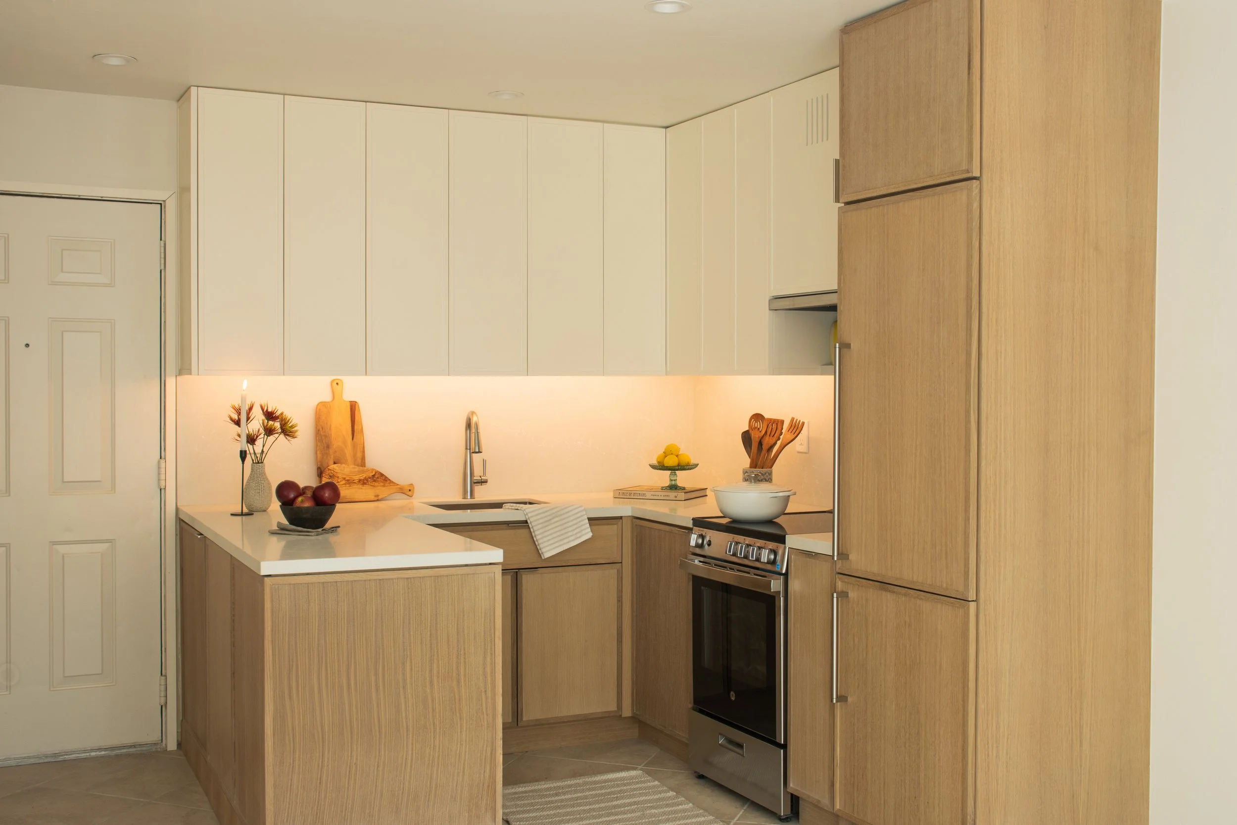Modern kitchen with light wood cabinets, white countertops, a stove, and decor including fruit, a candle, and kitchen utensils.