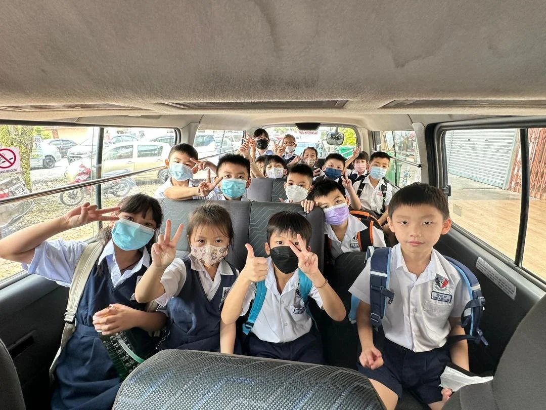 Group of students wearing school uniforms and face masks sitting inside a van, some making peace signs, with parking lot visible through the windows.
