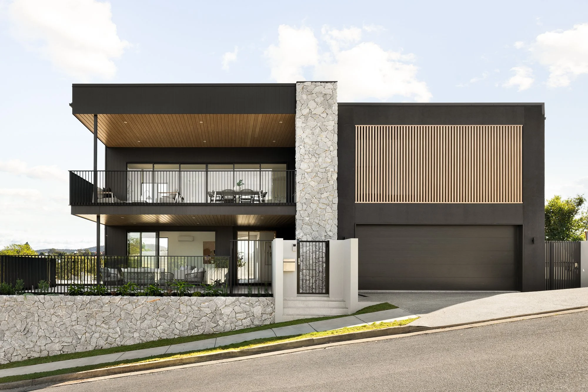 Modern two-story house with black exterior, stone accent wall, wooden accents, multiple balconies, and a garage on a slight hill.