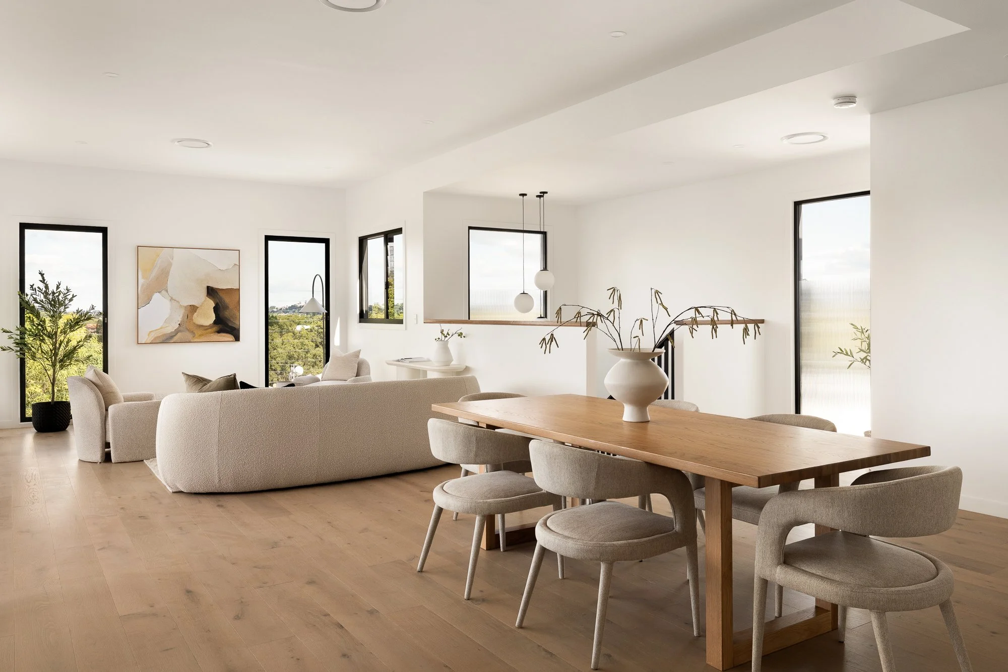 Modern, minimalist living room and dining area with beige furniture, wooden dining table, white walls, and large windows showing greenery outside.