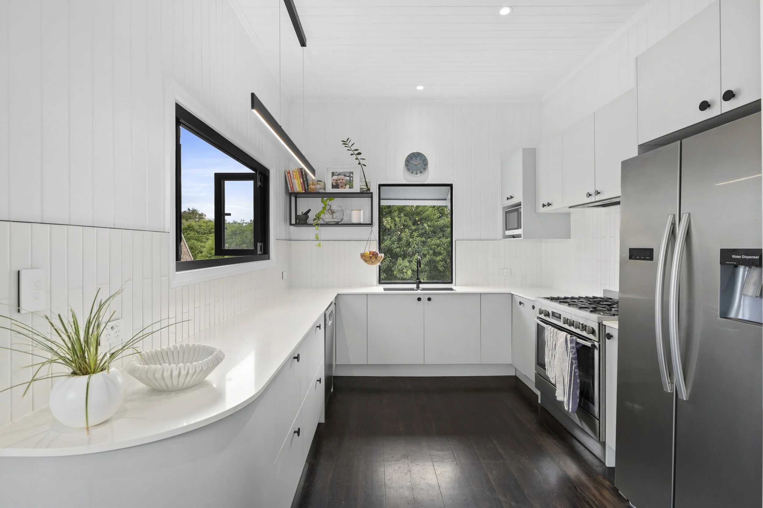 Modern white kitchen with black window frames, dark hardwood floors, white cabinets, and stainless steel appliances, including a refrigerator and oven.