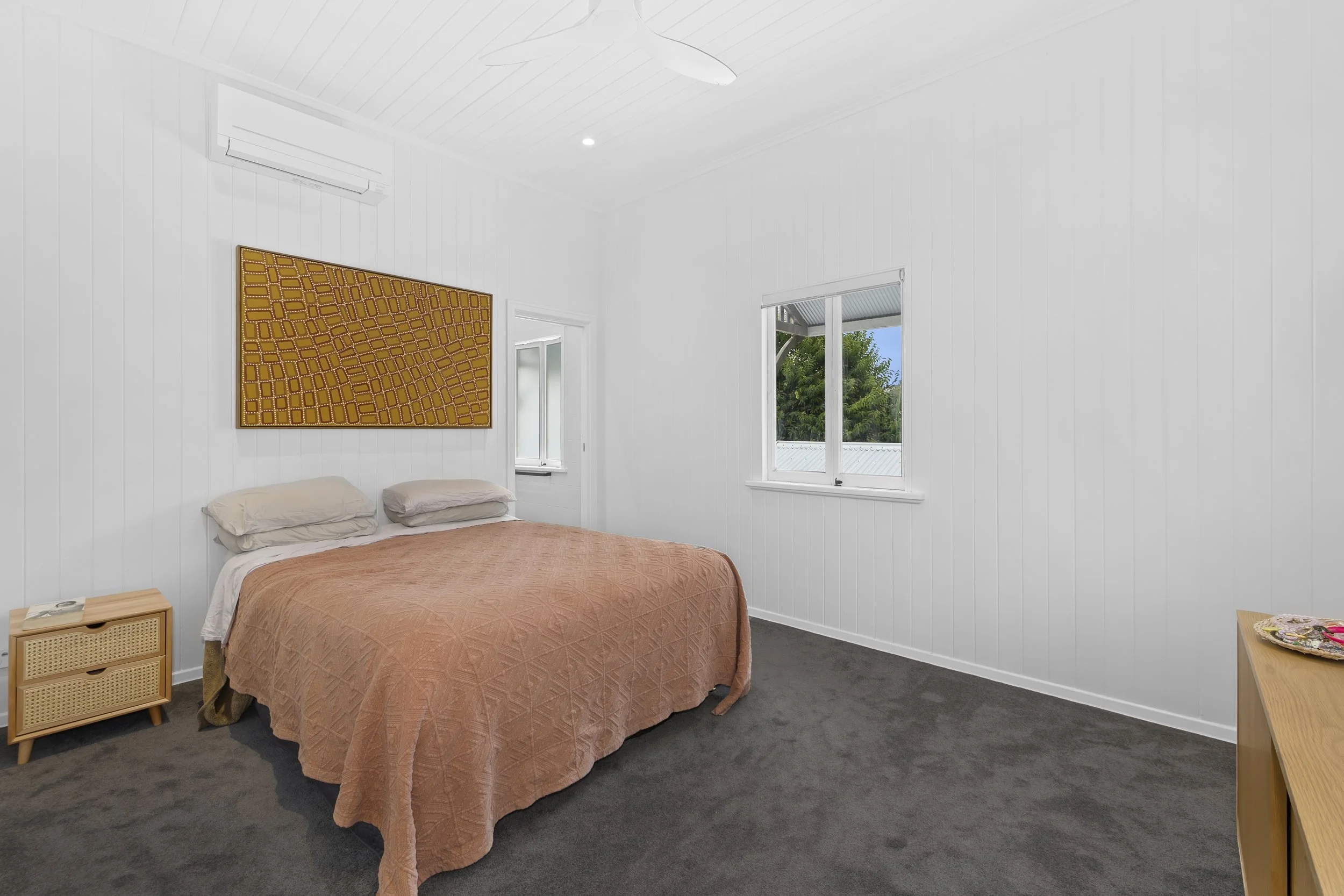 A bedroom with white paneled walls, two windows showing greenery outside, and a bed with a peach-colored quilt and two pillows. There is a small wooden nightstand with a drawer to the left of the bed and a wooden dresser with jewelry on the right. A