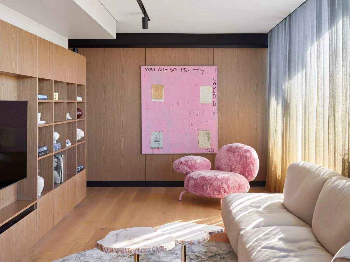 Modern living room with a beige sofa, pink furry chair, wooden floor, wooden wall with pink artwork, and large window with curtains.