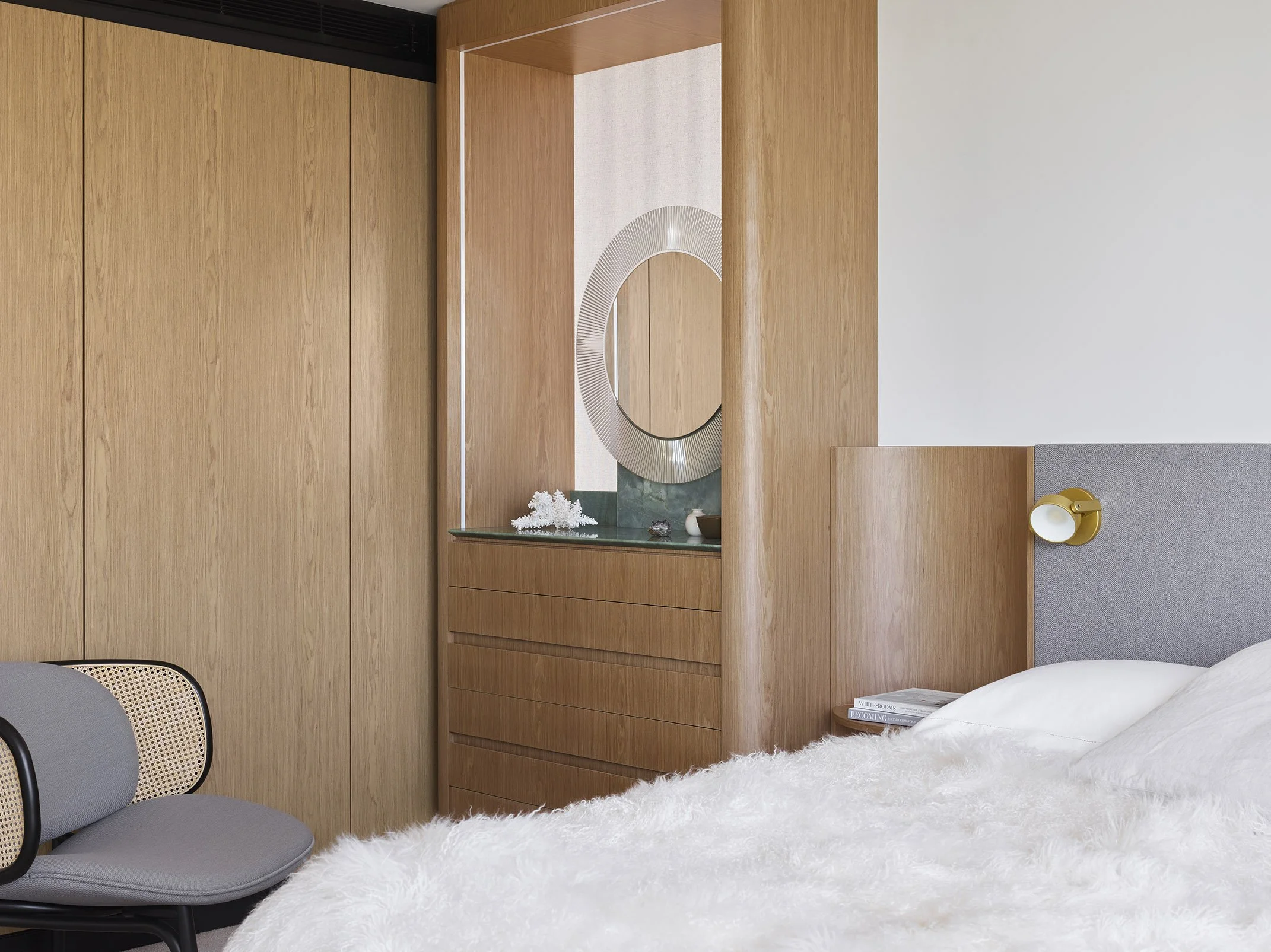 Modern bedroom with wooden paneling, a gray upholstered headboard, a gold wall-mounted reading light, a white soft blanket, a bedside table with a stack of books, a circular mirror, and decorative items on a dresser.