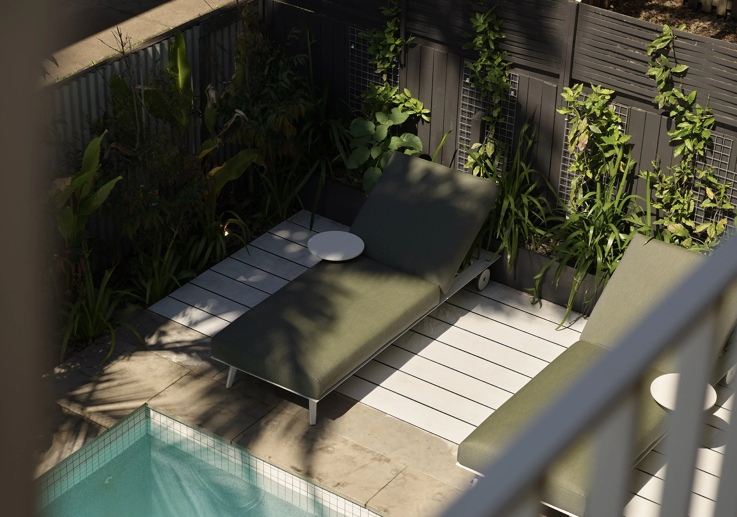 A small private outdoor patio with a swimming pool, two green lounge chairs, and lush green plants along a wooden fence.