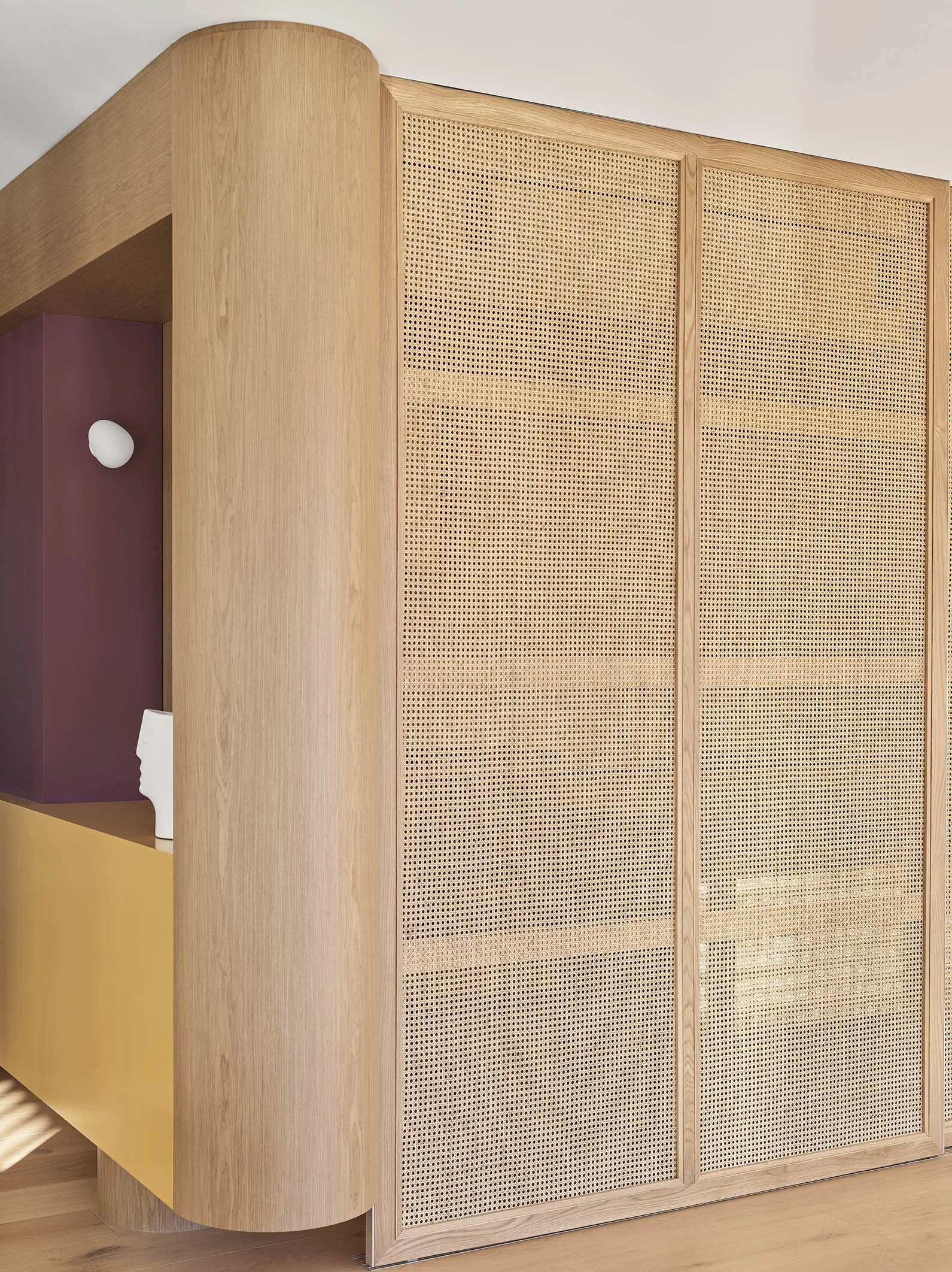 Close-up of a large wooden cabinet with round edges and perforated sliding doors, next to a colorful purple and yellow wall with decorative objects, in a modern interior.