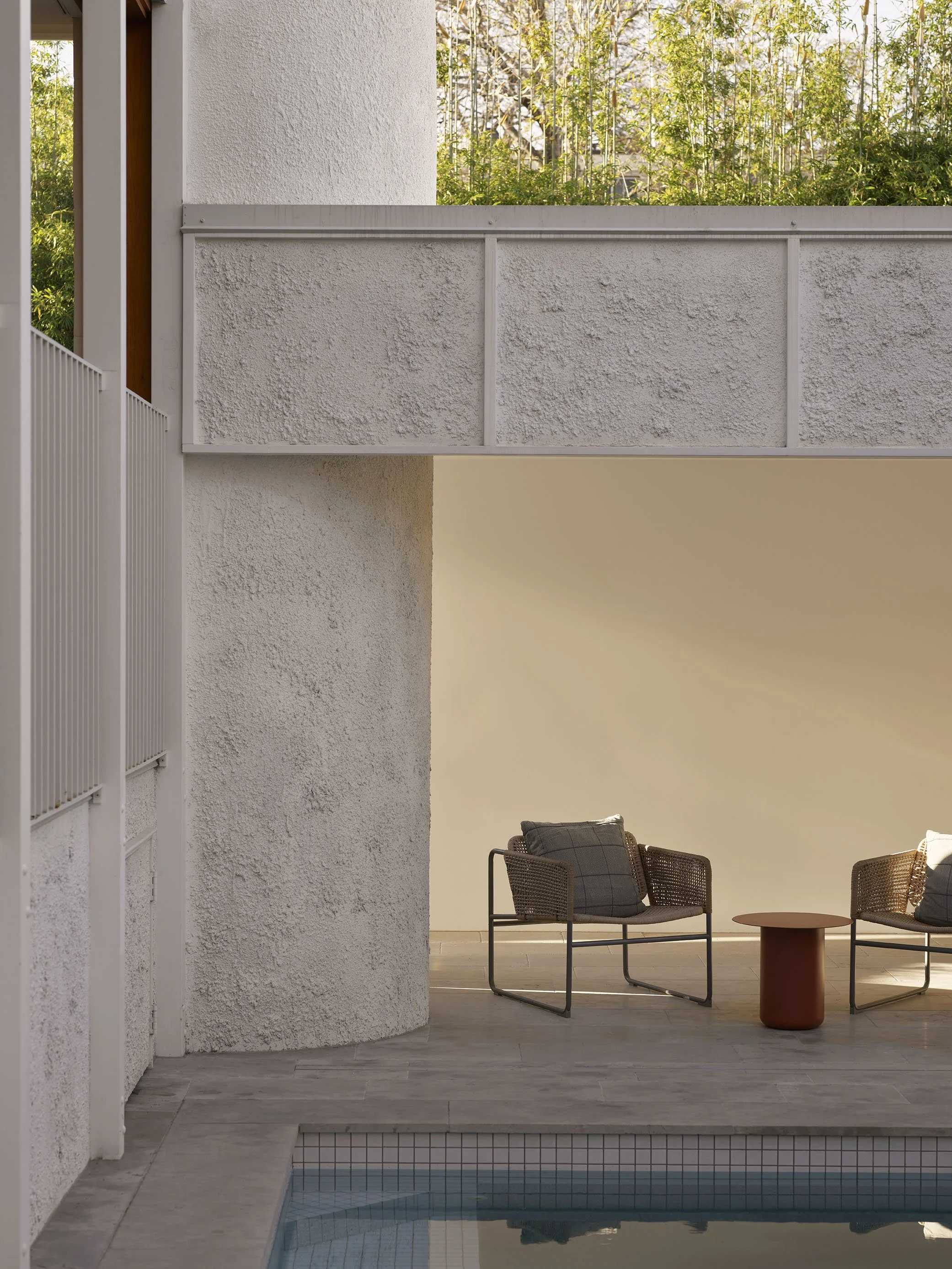 Modern outdoor patio with two wicker chairs and a small round table next to a swimming pool, with a textured white wall and greenery in the background.