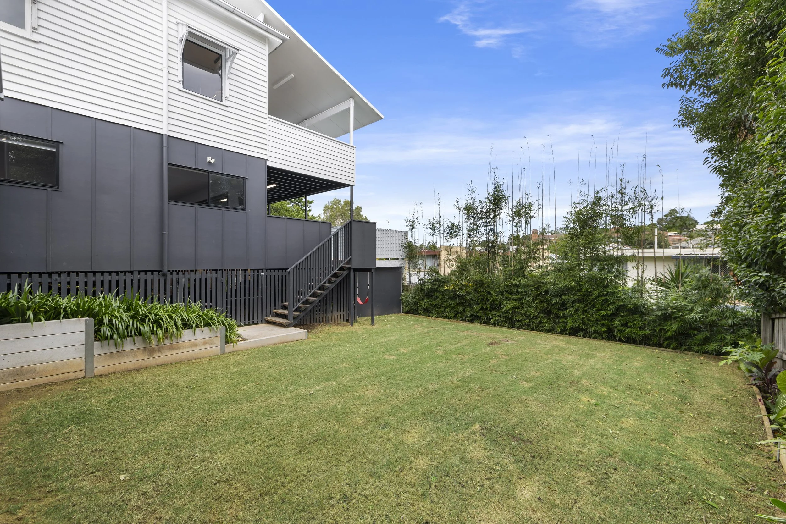 Backyard with green grass, wooden stairs leading to a covered balcony, and a large hedge of trees on the right. The house has white and gray siding and large windows.