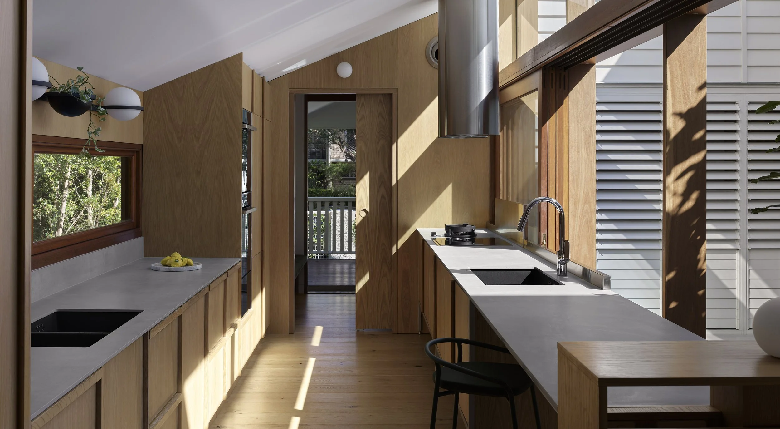 Modern kitchen with wooden cabinets, large windows, and a view of greenery outside. Sunlight streams through the windows, illuminating the countertops and floor.