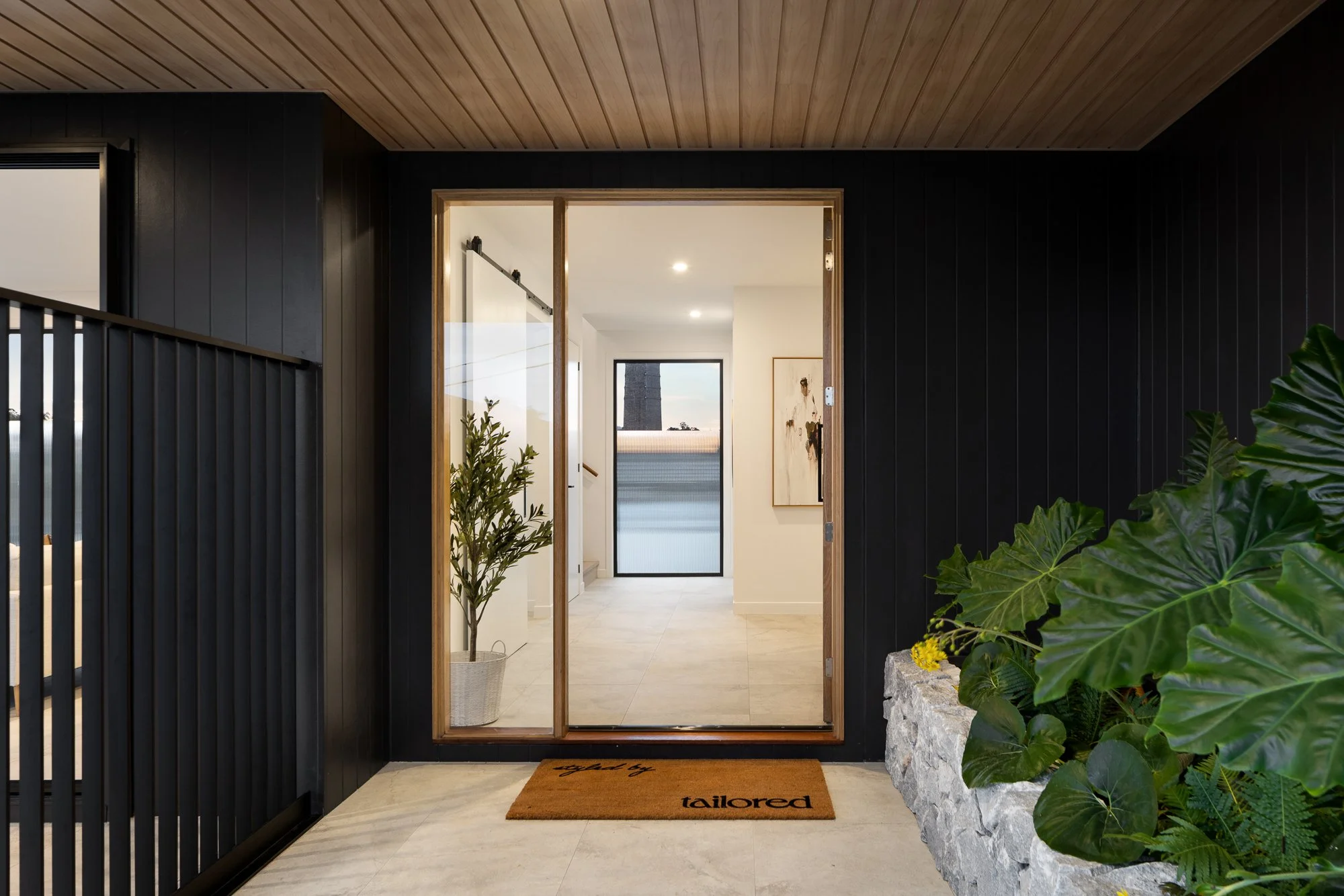 Entrance of a modern home with black walls, wooden ceiling, and a potted plant, with a doormat reading 'tailored' in front of a glass door leading inside.