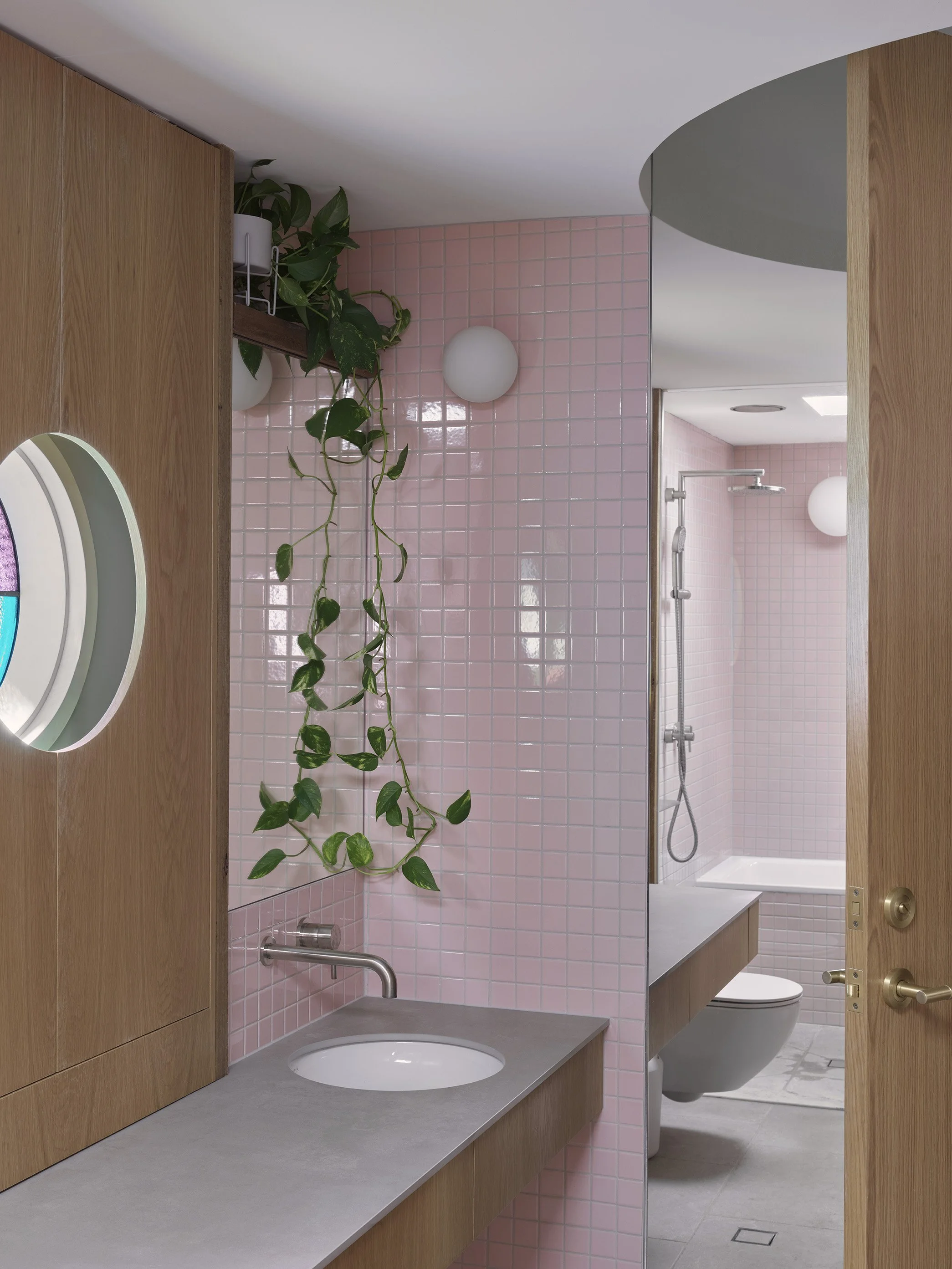 A bathroom with pink tiled walls, a wooden vanity with a concrete countertop and round mirror, a hanging plant, and a separate shower area with pink tiled walls and a white bathtub.