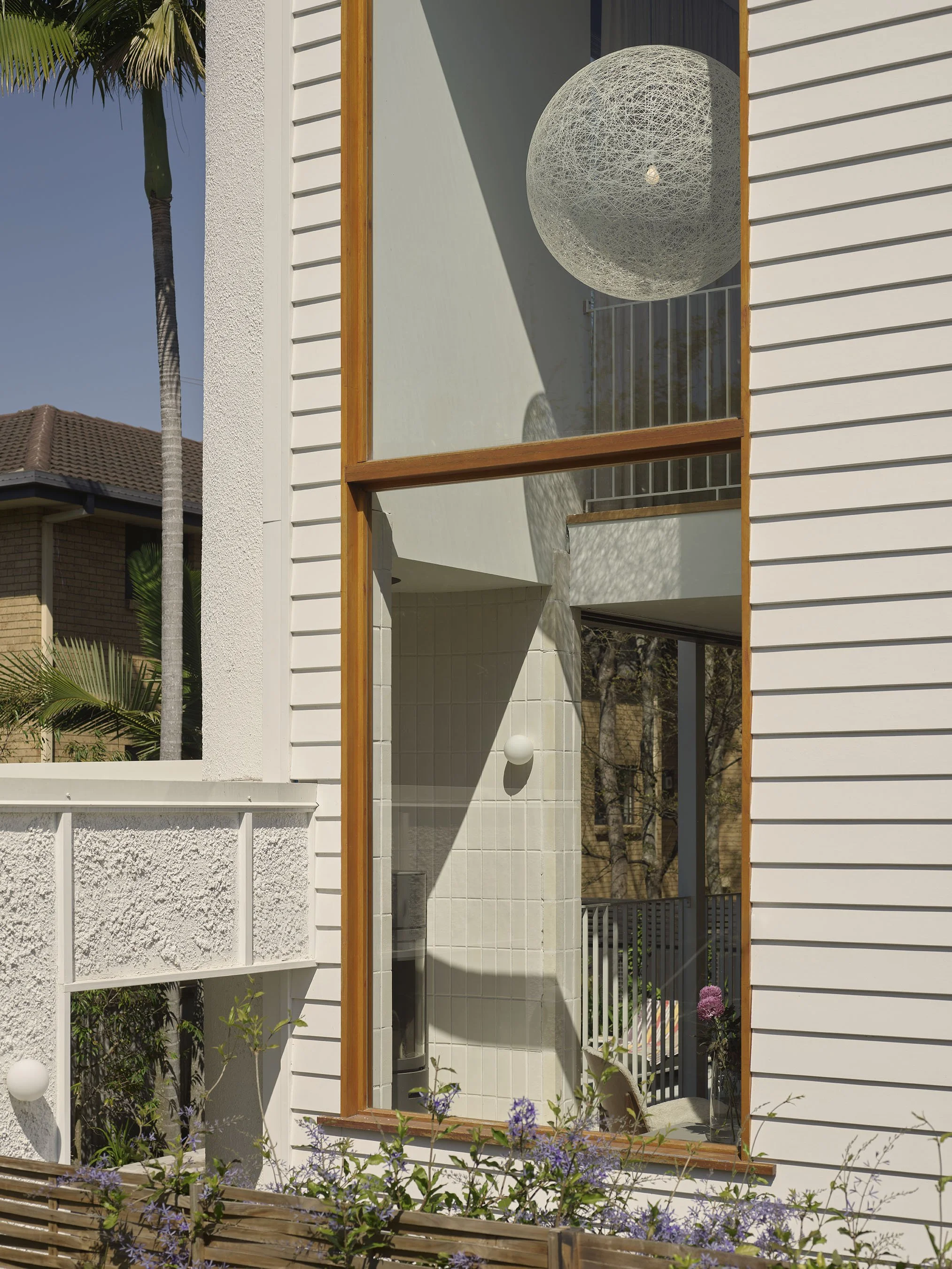 Close-up of a modern house exterior with white siding, large window showing interior decor, including a spherical light fixture and hanging light. There are plants and trees around the house, including a tall palm tree.