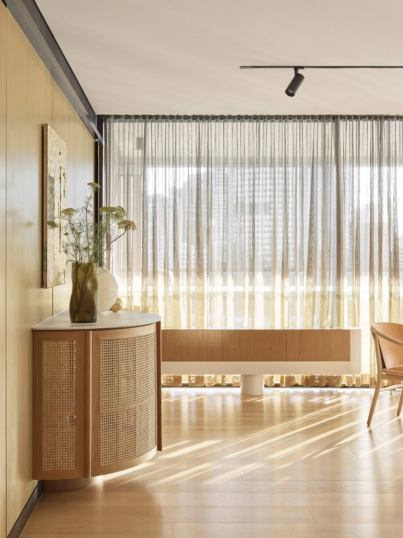 A bright, modern interior with light wooden flooring, a curved wooden cabinet with rattan doors, a vase with tall dried flowers on top, beige curtains allowing sunlight, and a pink chair with a rounded back to the right.