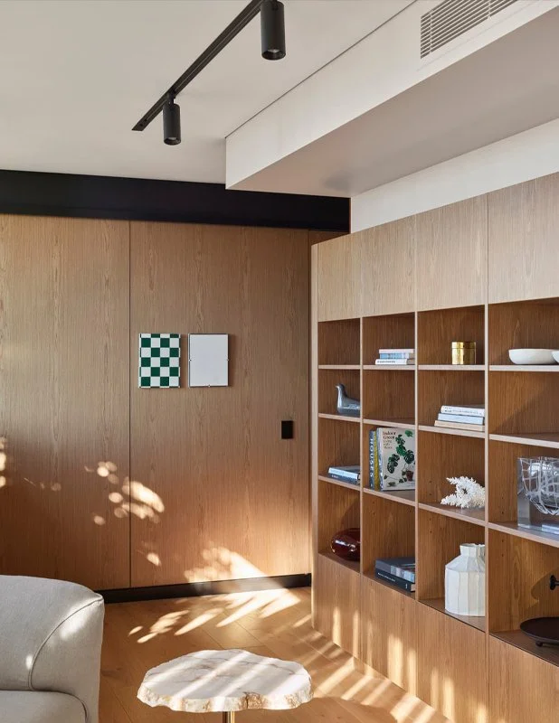 A modern interior corner with wooden shelving filled with books and decorative items, sunlight casting shadow patterns on the wall and floor, beige sofa partially visible, and a small white marble side table.