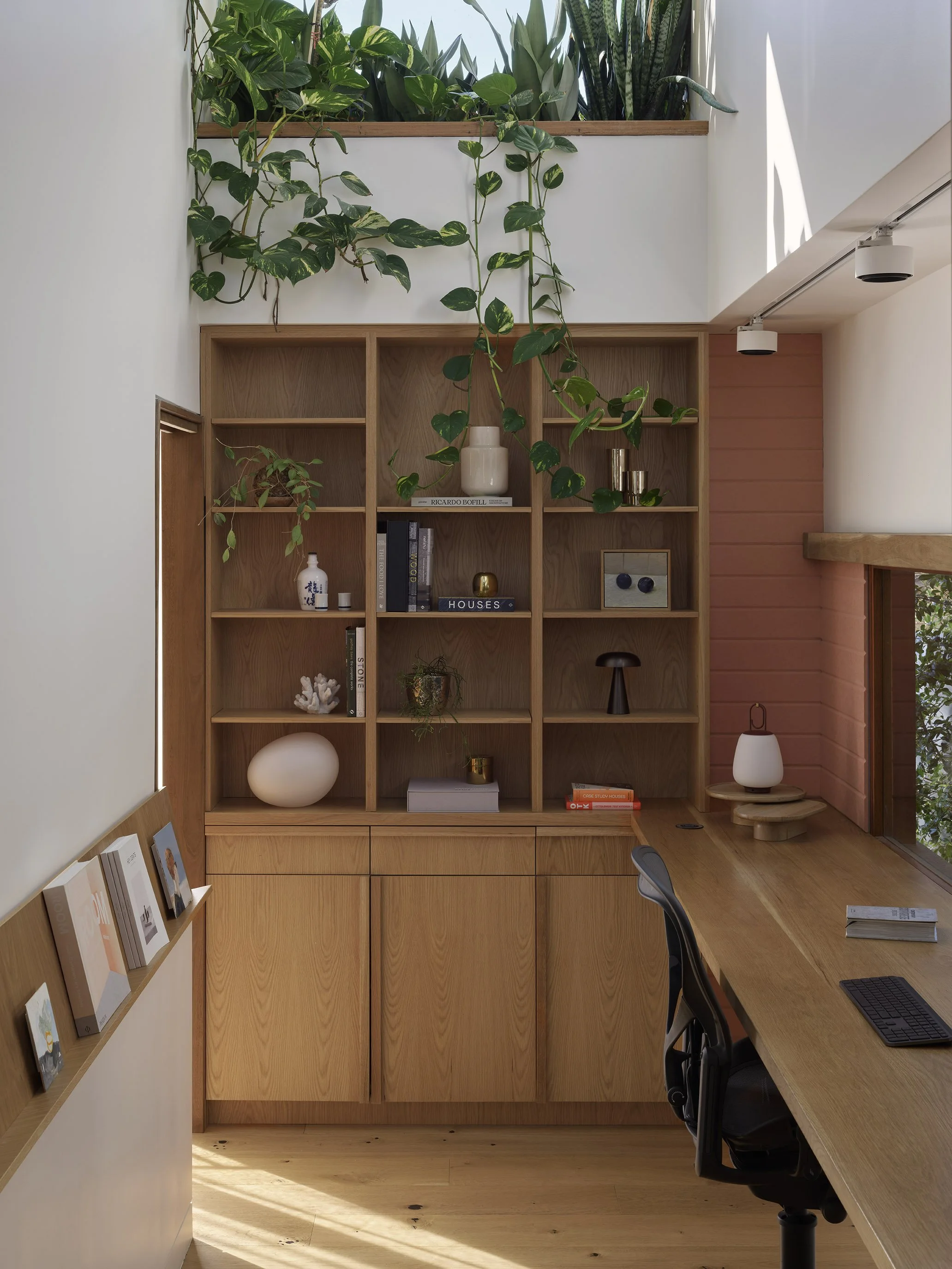 A wooden office desk with a black office chair, books, a keyboard, and a notepad. Behind the desk is a wooden shelving unit with various decorative items, books, and plants. Green leafy plants grow above the shelf, and sunlight comes through a nearby