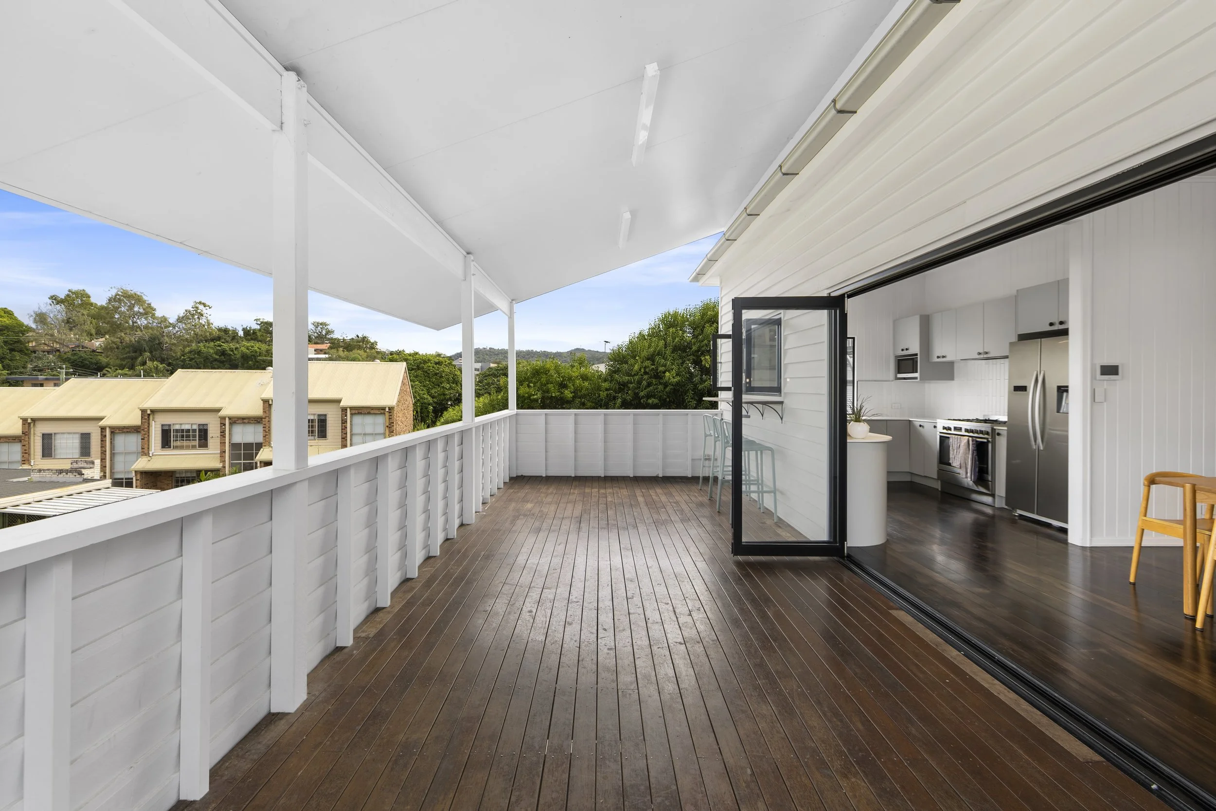 Empty balcony with wooden floor, white railing, and open sliding glass door leading to a modern white kitchen.