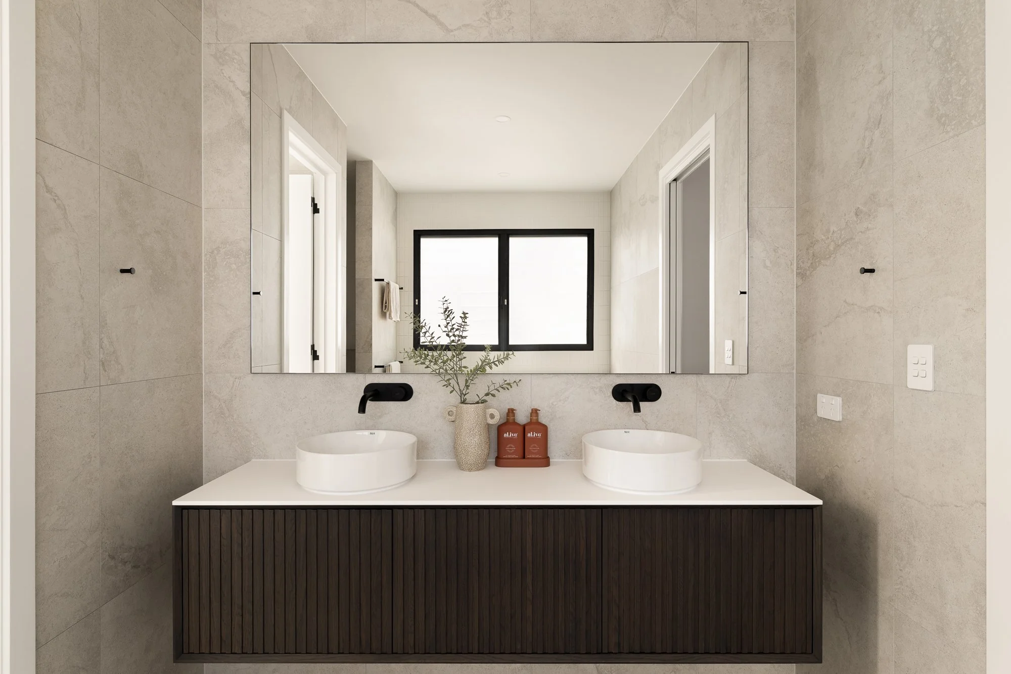 Modern bathroom with double vanity, large mirror, and two vessel sinks, beige tiled walls, and a window.