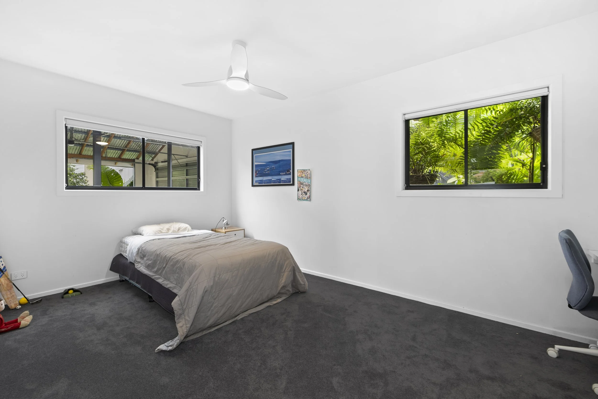 A simple bedroom with white walls, a bed with beige bedding, a small nightstand, two windows showing green foliage outside, a ceiling fan, a framed picture, and a gray office chair.