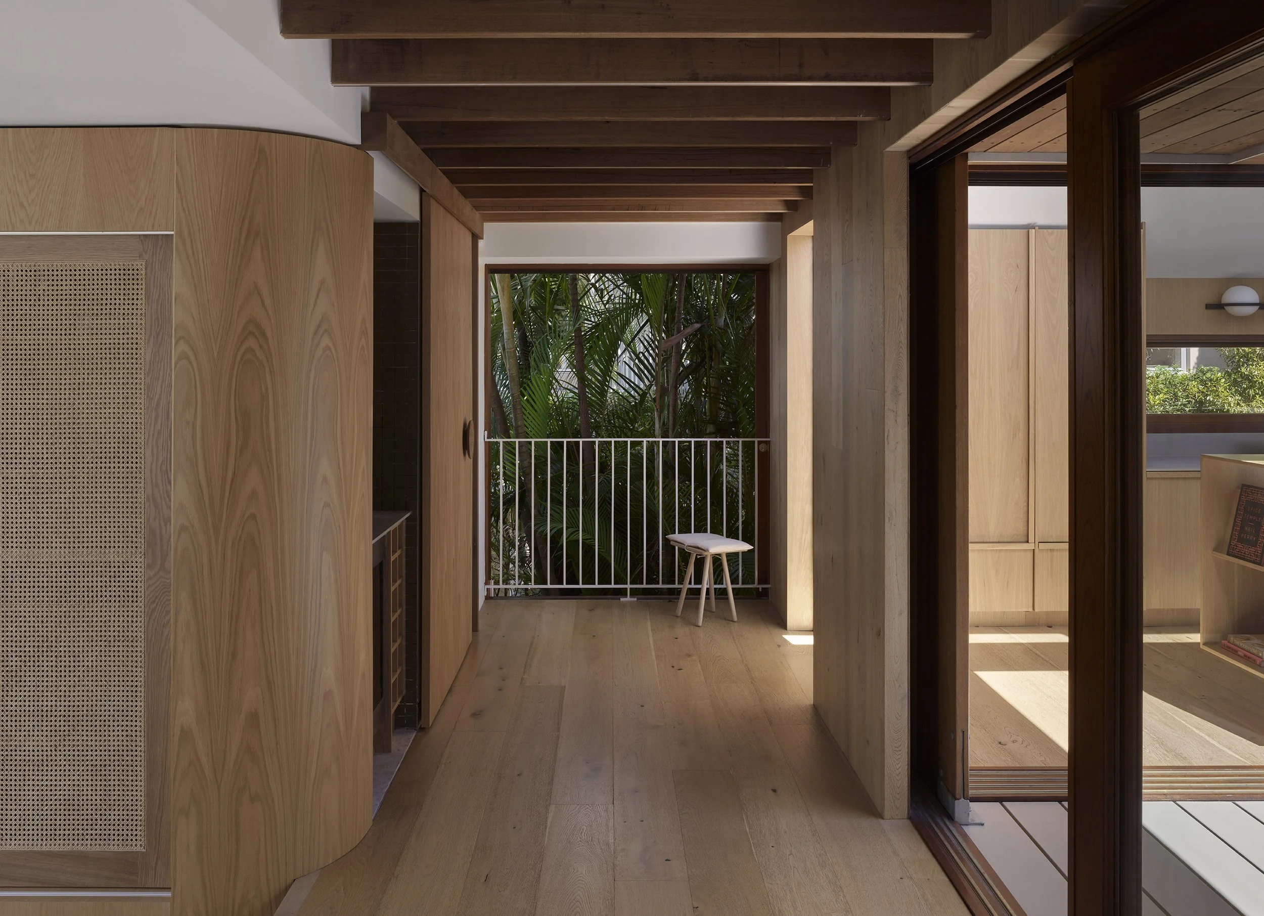 Interior view of a room with wooden walls, flooring, and ceiling beams. A balcony with greenery is visible through the open sliding glass door, with a small white stool near the balcony railing.