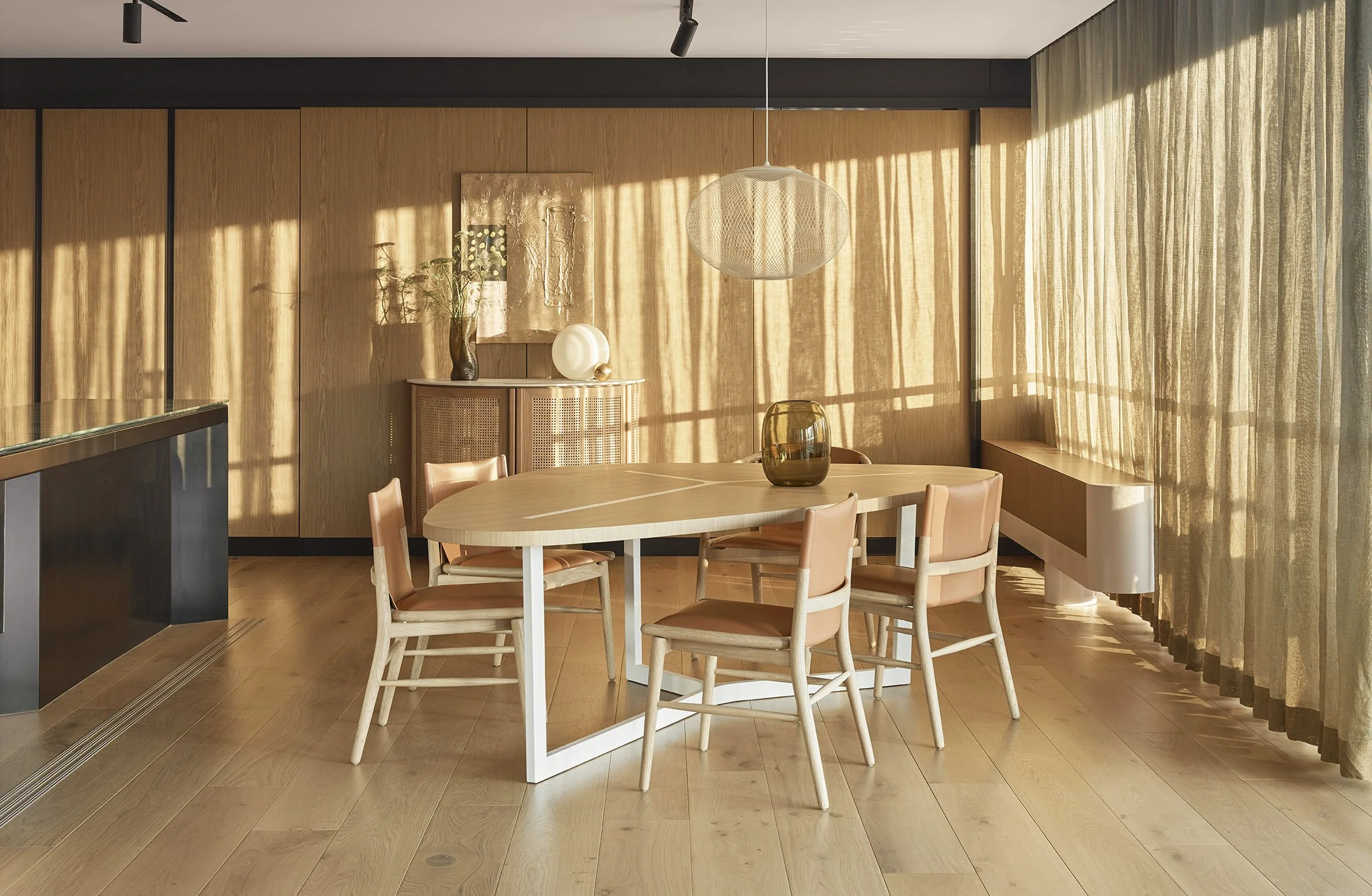 Modern dining room with wooden walls, a round table with four chairs, sheer curtains, and minimalist decorative items illuminated by natural sunlight.
