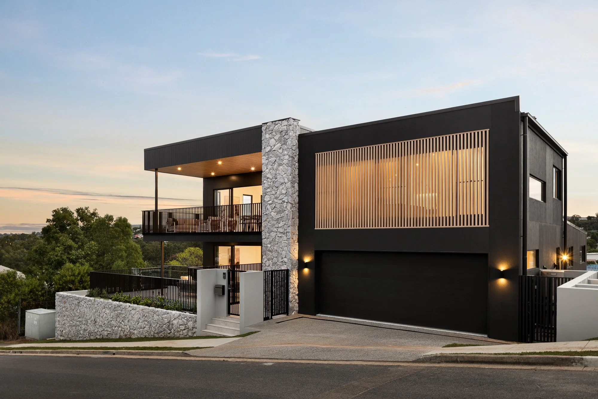 Modern two-story house with black exterior, stone accents, large balcony, and warm lighting, situated on a slightly inclined street during sunset.