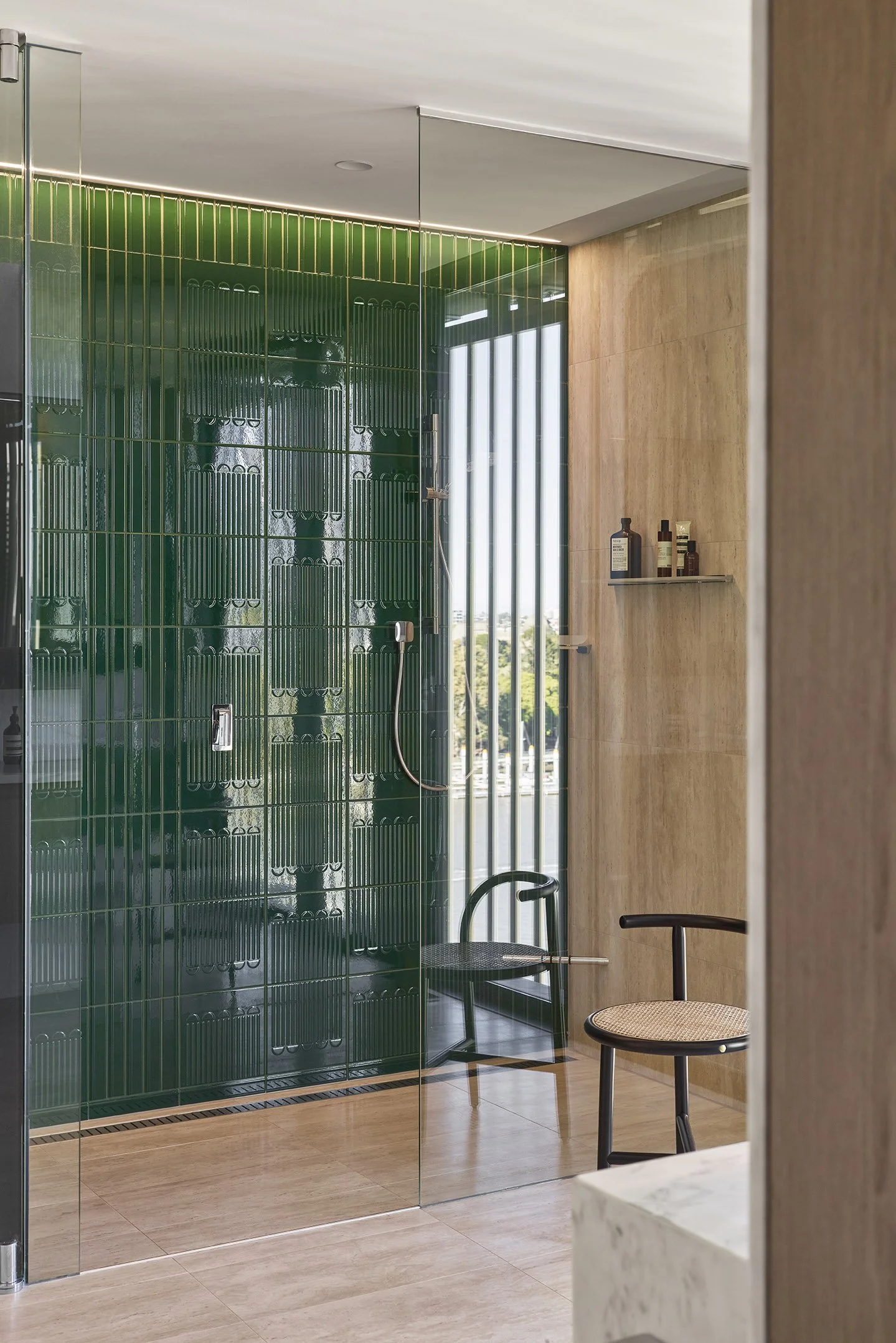 Modern shower with green tiled wall, glass door, and two black chairs nearby.