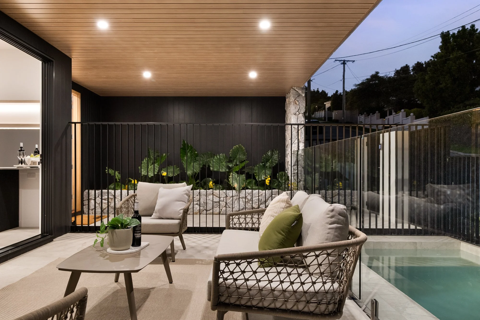 Covered outdoor patio with seating area, potted plant, and view of a backyard pool at dusk.