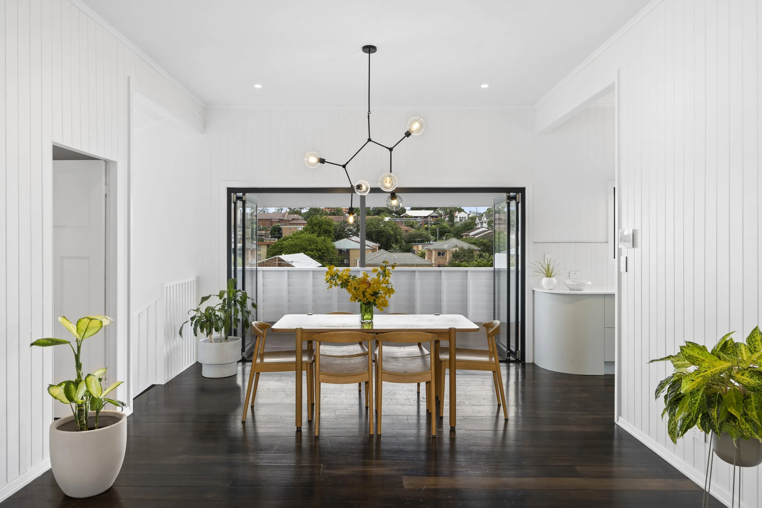 Bright, modern dining room with white walls, dark wooden floors, and large open doors leading to a balcony with views of a suburban neighborhood. There is a wooden dining table with six chairs and a vase of yellow flowers, potted plants on the sides,