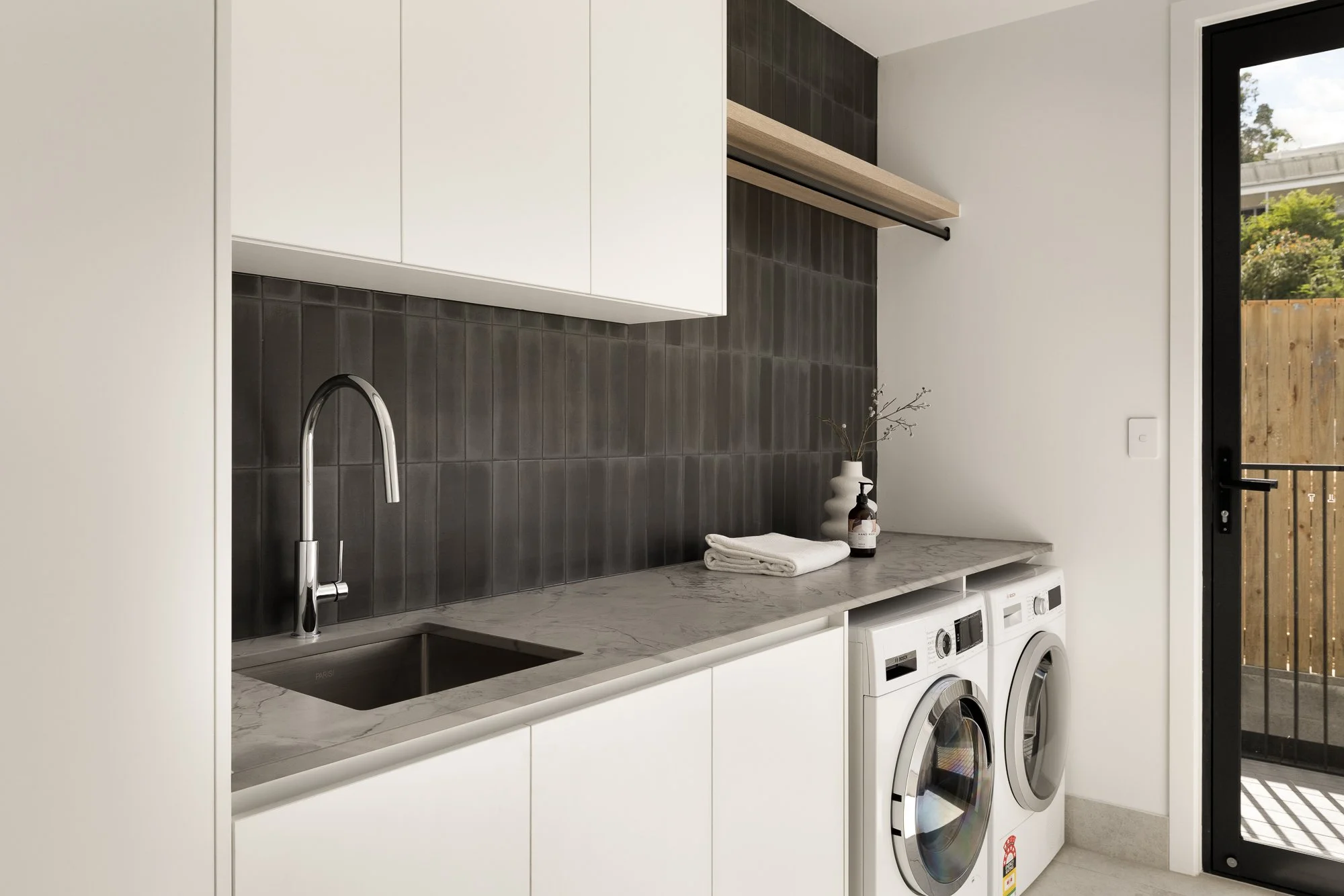 Laundry room with white cabinets, a marble countertop, a black backsplash, a stainless steel sink, a faucet, a folded towel, a soap dispenser, a vase with branches, a washer and dryer, and a glass door leading outside.