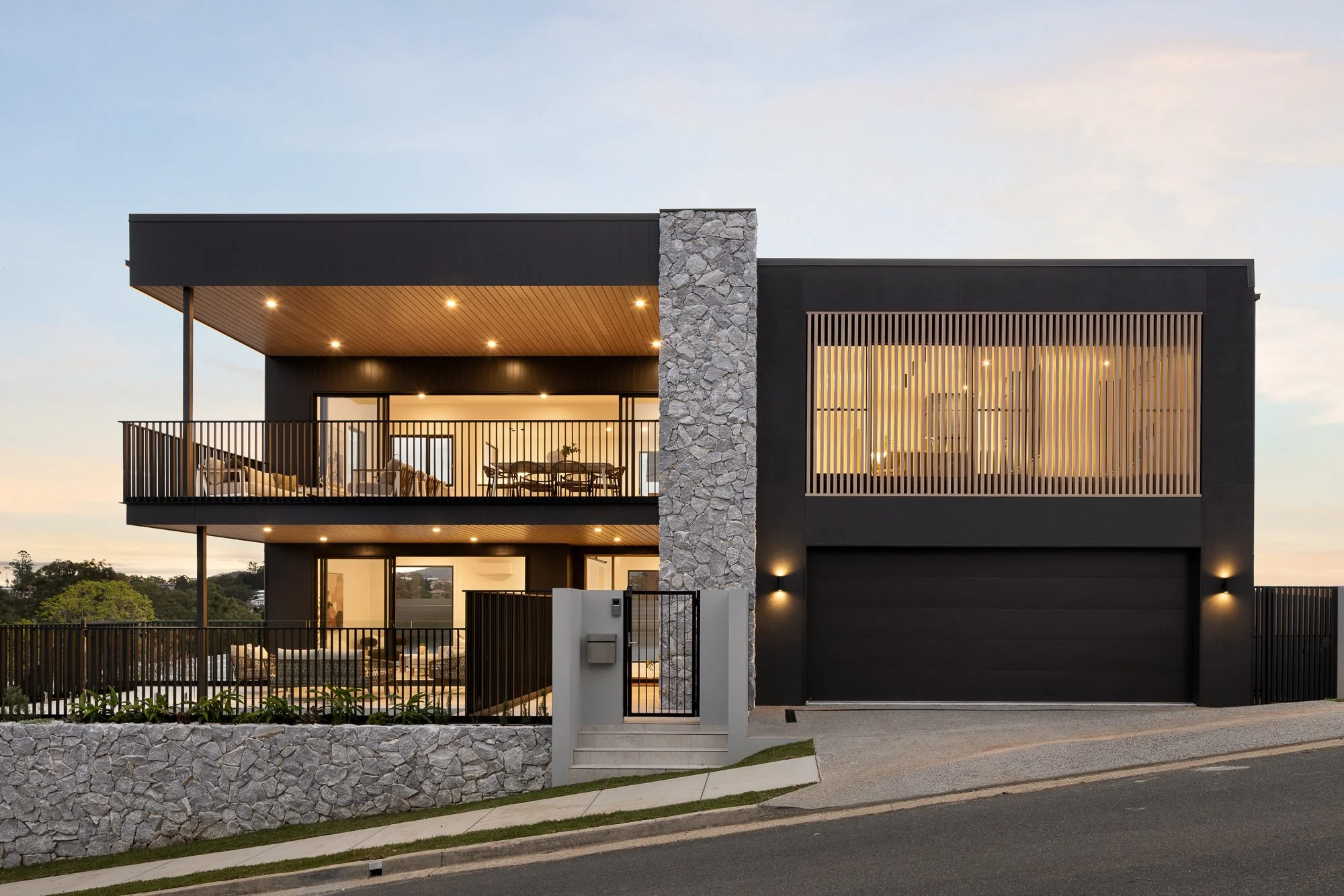 Modern two-story house with black exterior, stone accents, large windows, and balconies, illuminated at dusk.