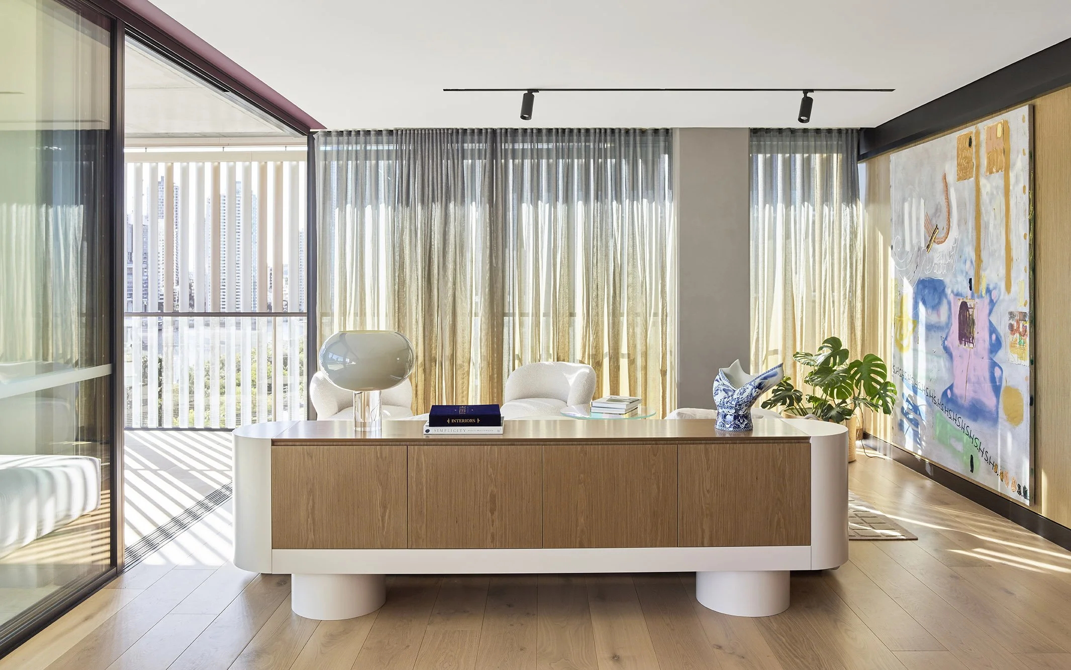 Modern office with wooden desk, white and blue decorative objects, a plant, and large floor-to-ceiling windows with curtains, overlooking city buildings.