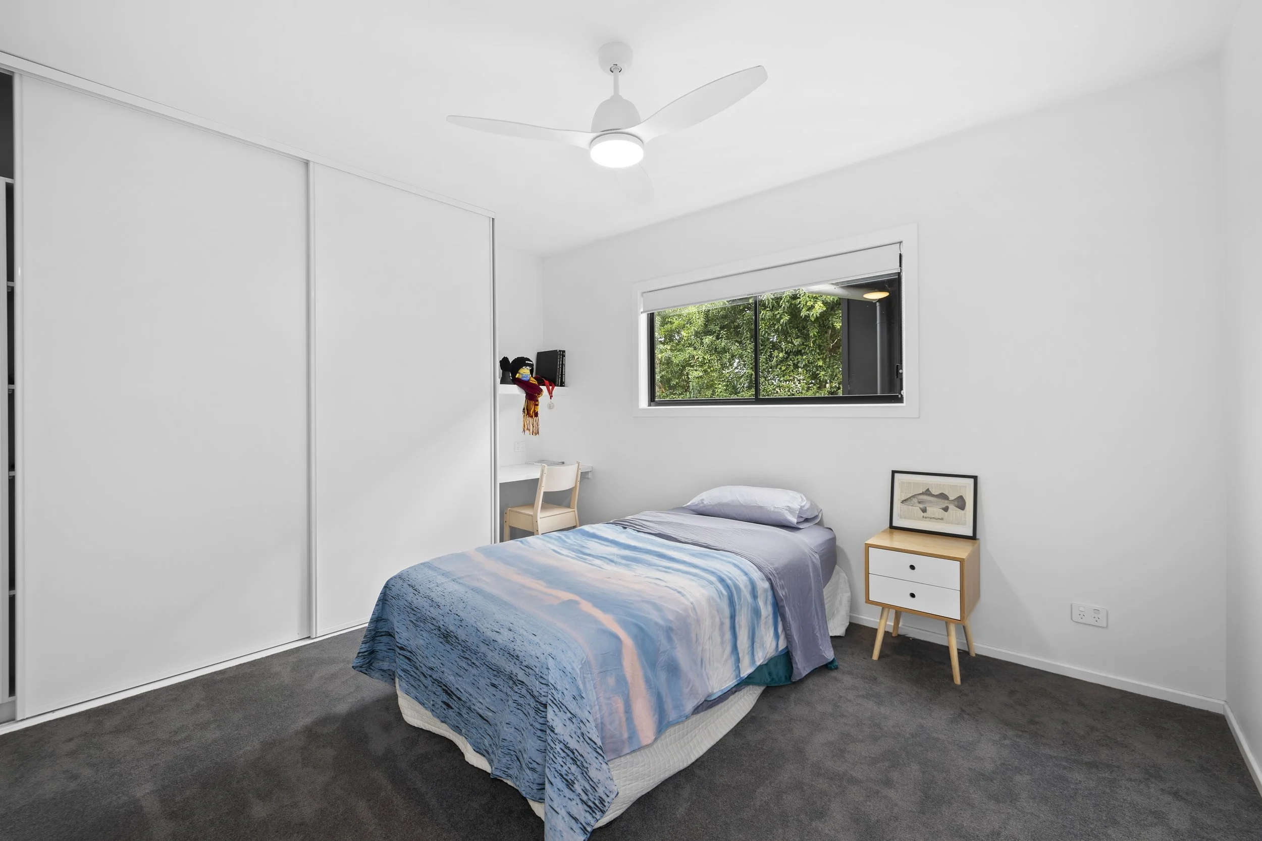 Simple bedroom with a single bed, white walls, gray carpet, small white nightstand with a framed fish print, window showing greenery outside, ceiling fan, and a white desk with a chair and plush toy in the corner
