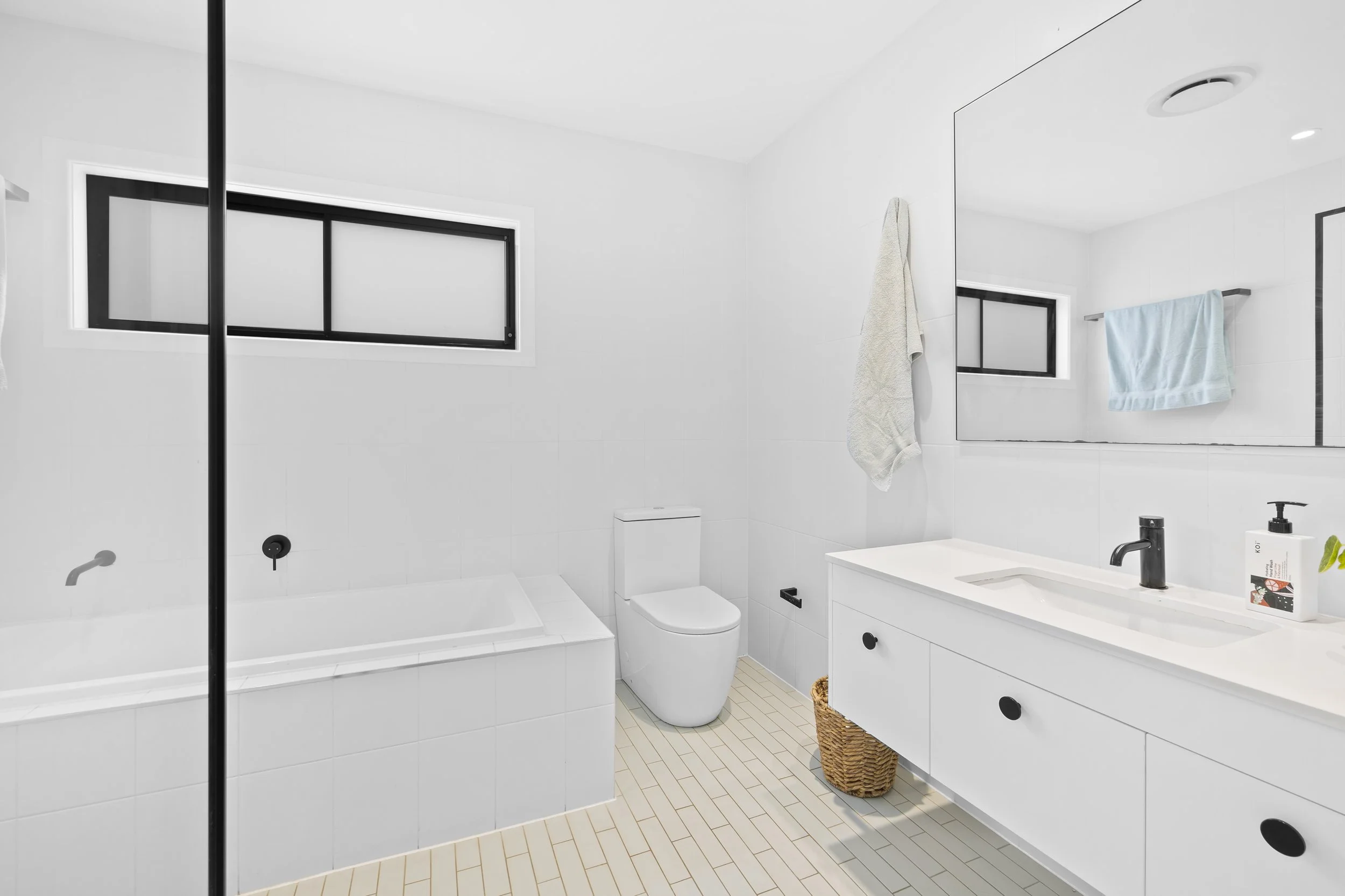 Modern white bathroom with a bathtub, toilet, large mirror, black fixtures, and a window with black framing