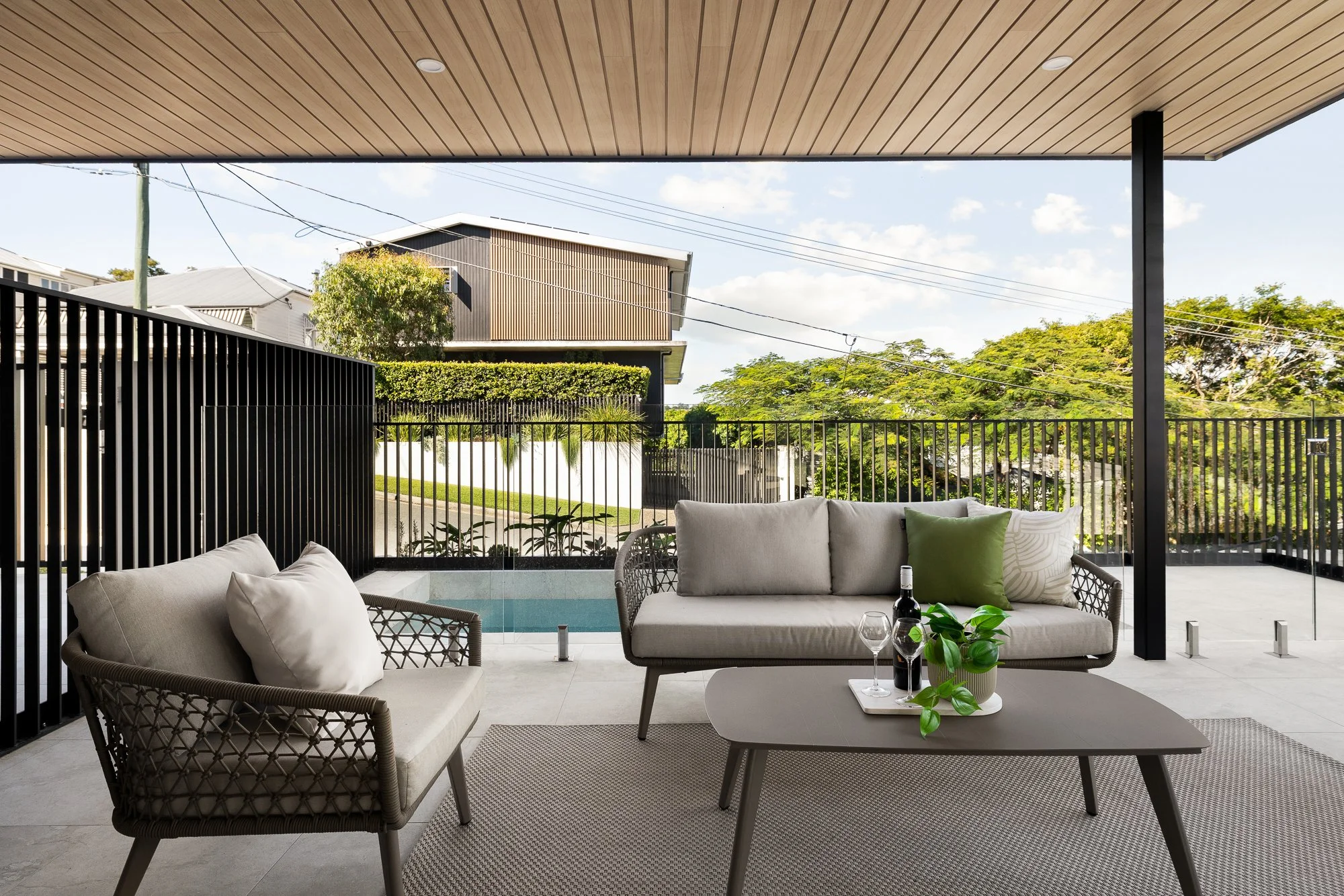 Modern outdoor patio with beige cushioned seating, a small coffee table with a green plant and drinks, overlooking a backyard with trees and a house in the distance.