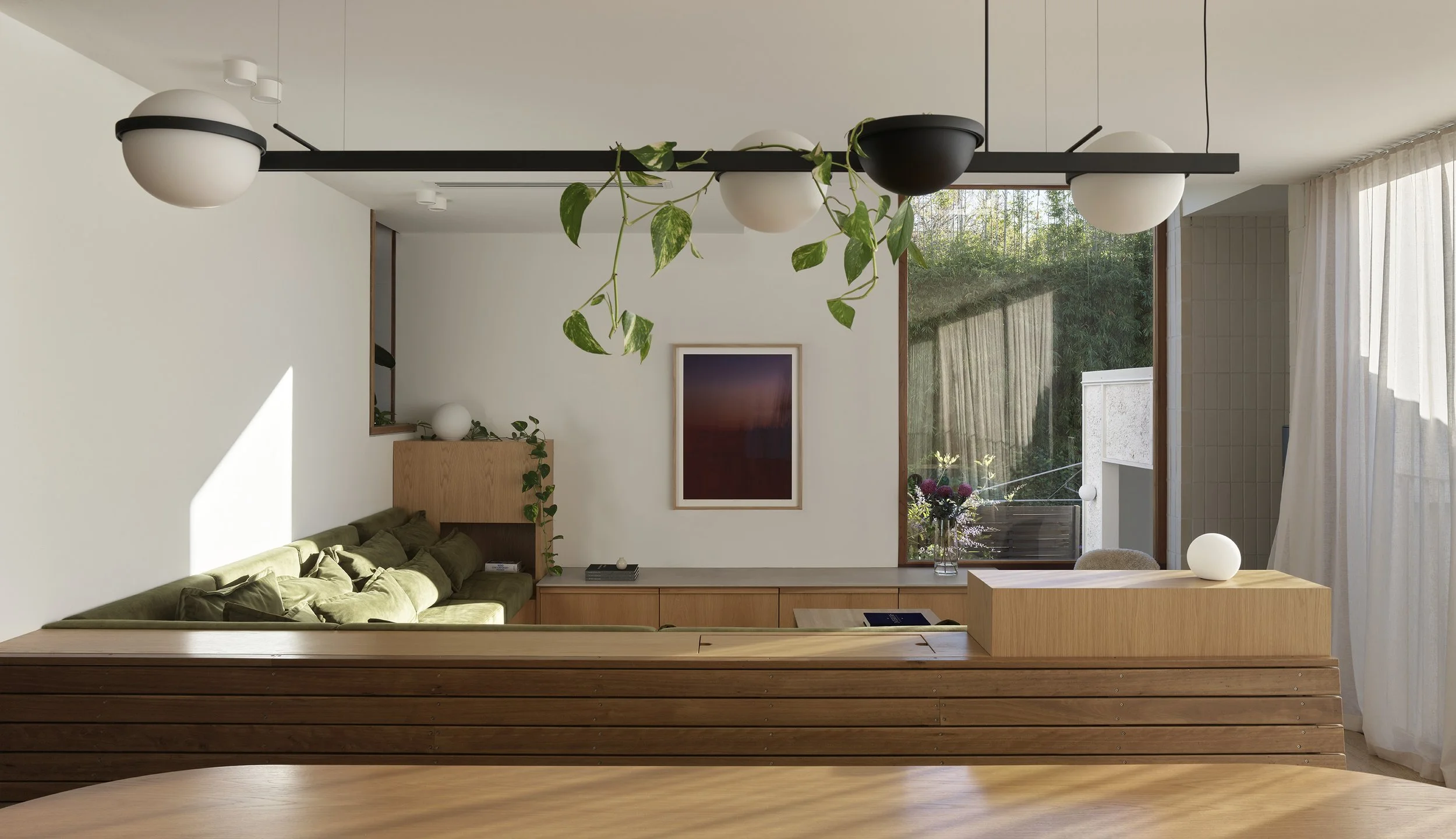 Modern living room with green couch, wooden shelf, and large window with sheer curtains, decorated with potted plants and flower vase.