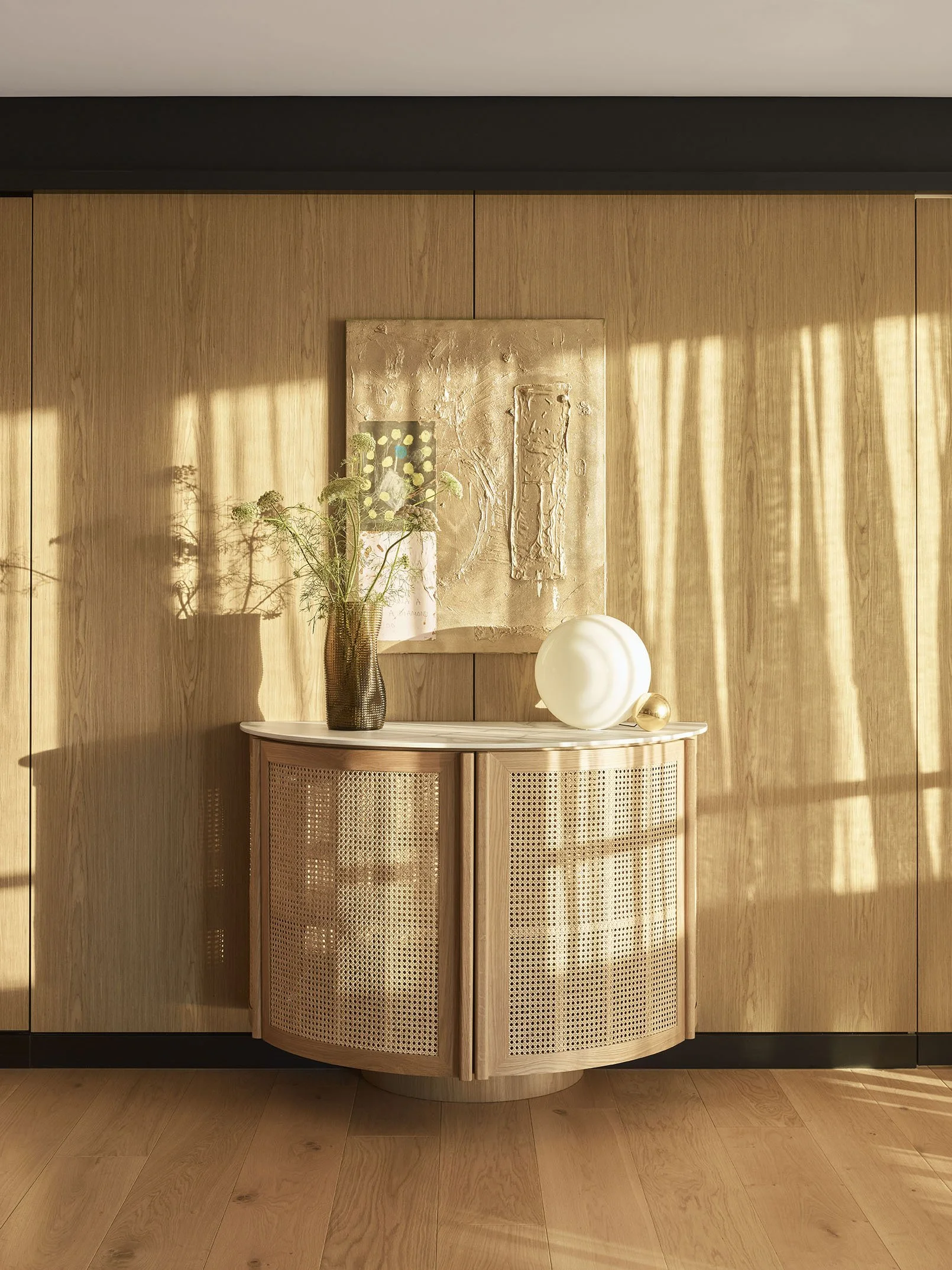 Wood-paneled wall with a decorative cabinet, a tall vase with green foliage, and two modern spherical sculptures, illuminated by warm sunlight.