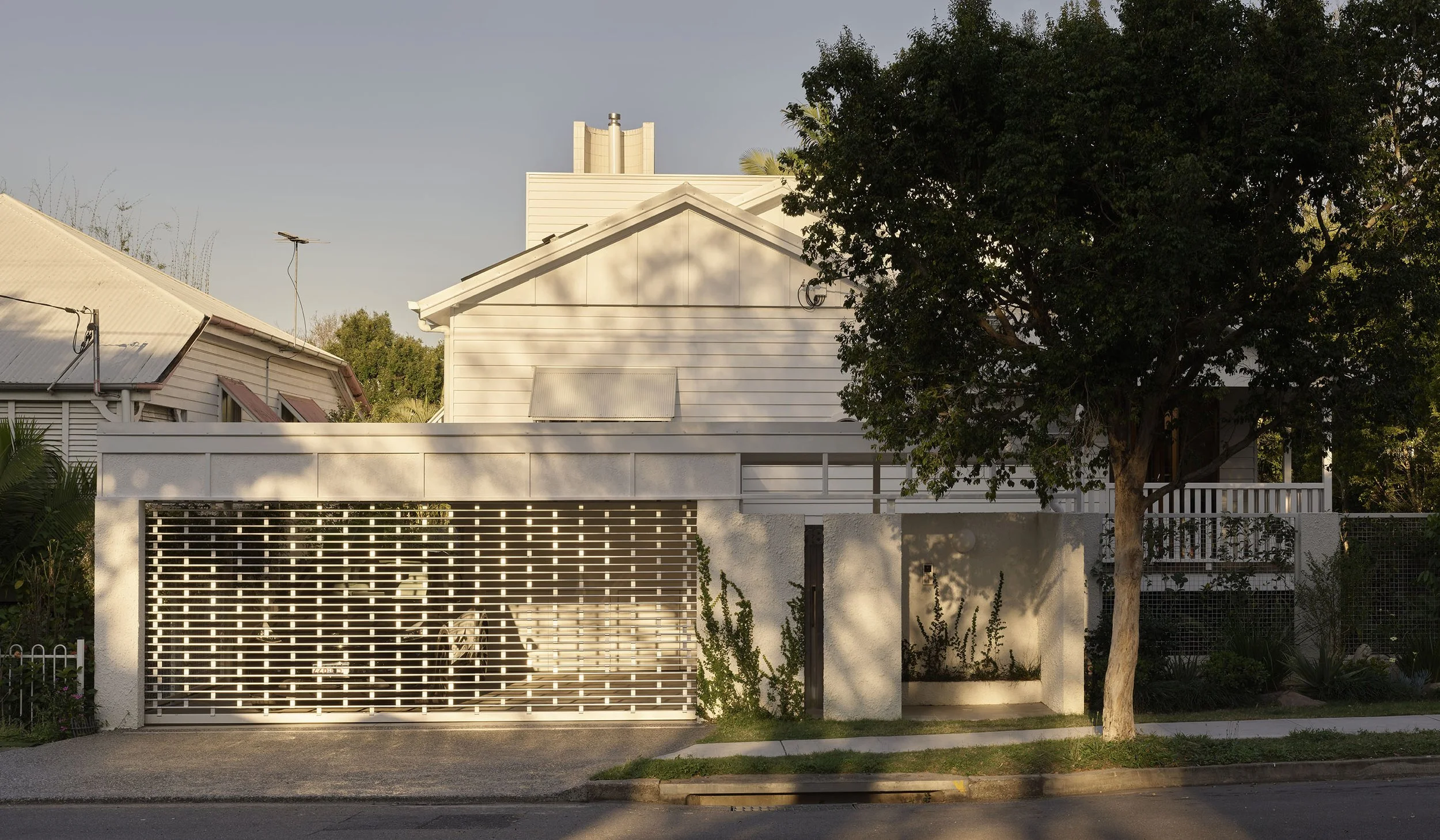 A white house with a gated entrance, a tree in front, and a sidewalk. The house has horizontal siding and a small balcony, with greenery and plants around.