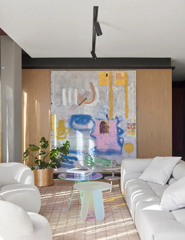 Modern living room with white sofas, colorful modern art painting on wooden wall, potted plant, and pastel-colored coffee tables with stacked books.