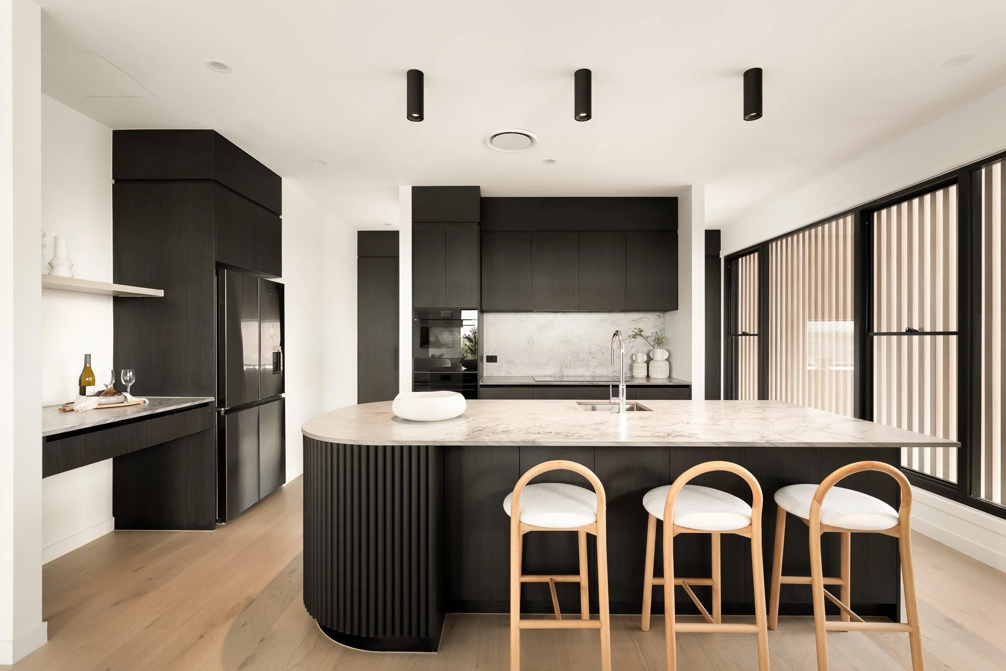 Modern kitchen with a large central island with a white marble countertop, three light wooden chairs, dark wood cabinetry, a stainless steel refrigerator, and large window with black frames and vertical blinds.