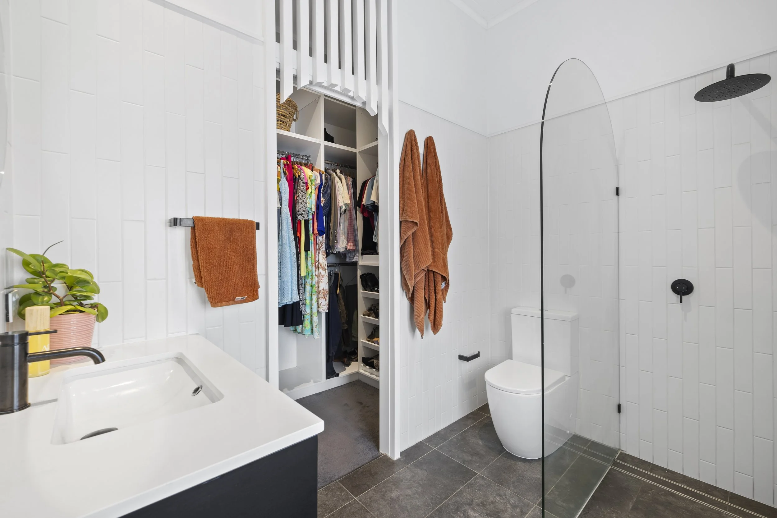 Bathroom with a walk-in closet, white tiled walls, a corner toilet, shower with a black rainfall showerhead, a black round control, and a thin glass divider. There are brown towels hanging and a small to medium plant on the white sink counter. The wa