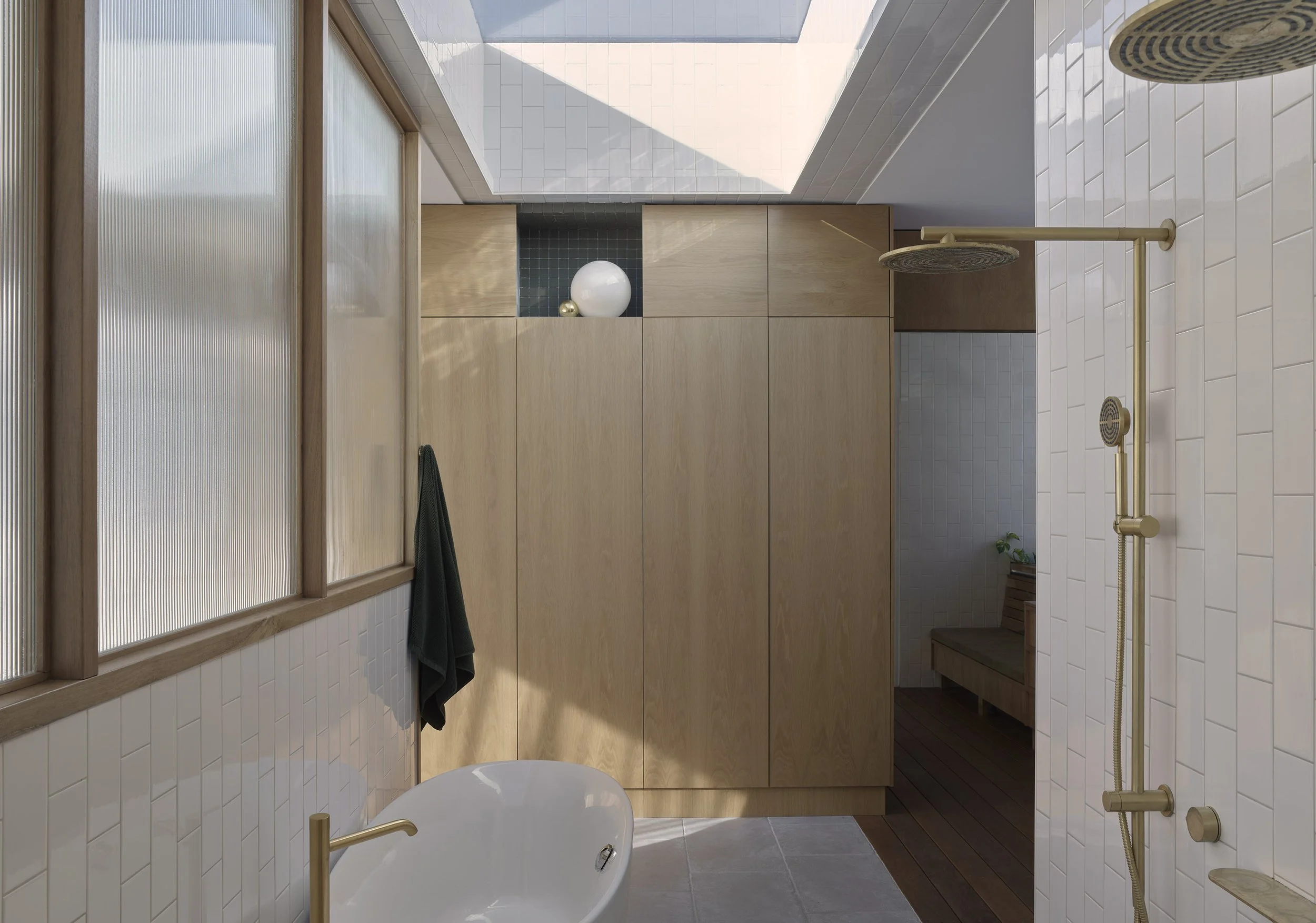 Modern bathroom with a white freestanding bathtub, a black towel hanging on wooden wall panels, a large window with frosted glass, a beige shower system, and wooden cabinets. Decorative items include white and gold balls and a green plant.