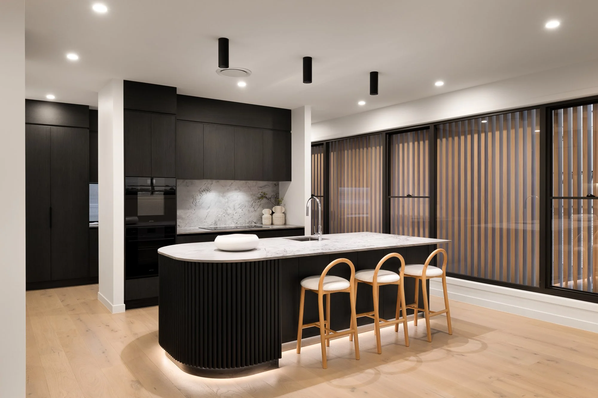 Modern kitchen with black cabinetry, a white marble island, wooden bar stools, floor-to-ceiling windows with wooden slats, and recessed ceiling lights.