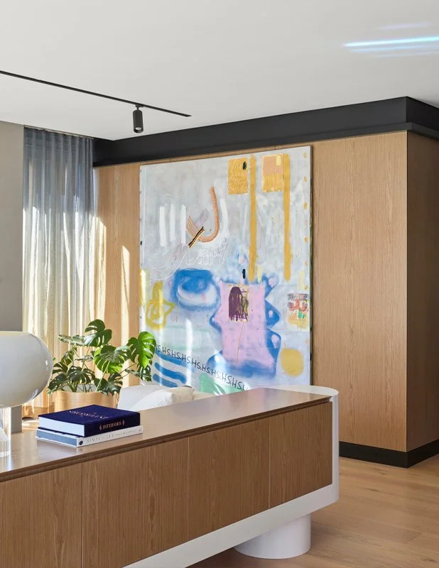 Modern interior room with large abstract painting on wood-paneled wall, green leafy plant, white sideboard with books and a white decorative object, sheer curtains, and ceiling spotlight.