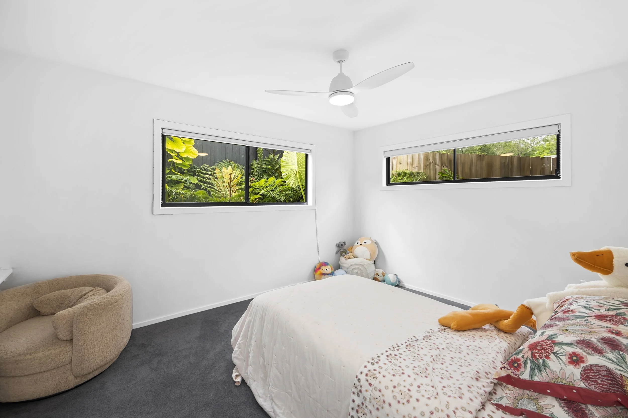 A minimalist bedroom with white walls, two horizontal windows showing green foliage outside, a ceiling fan, a beige pet bed, a bed with a floral-patterned pillow, plush toys including a duck and stuffed animals, and a dark carpet.