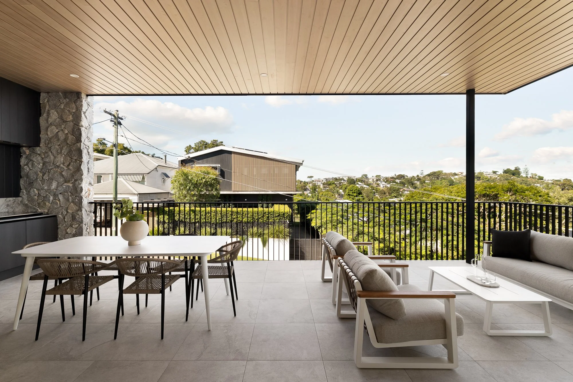 Modern balcony with a white dining table and six chairs, a beige sofa, and a view of trees and houses under a partly cloudy sky.