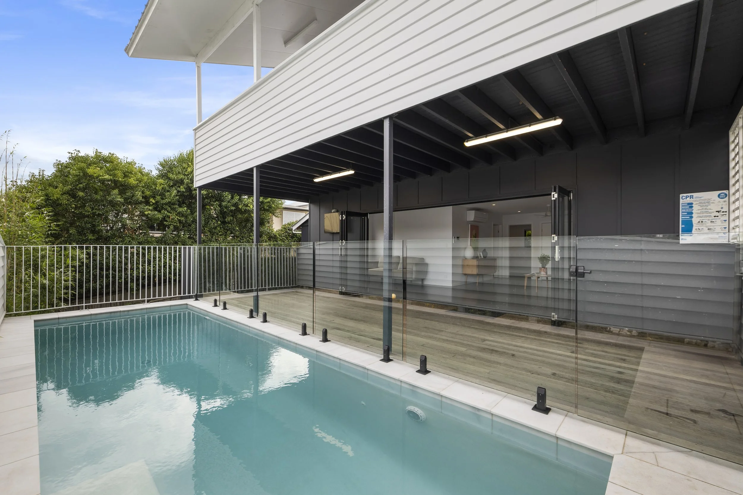 Modern backyard with a swimming pool enclosed by a glass fence, adjacent to a house with an open living area visible inside.