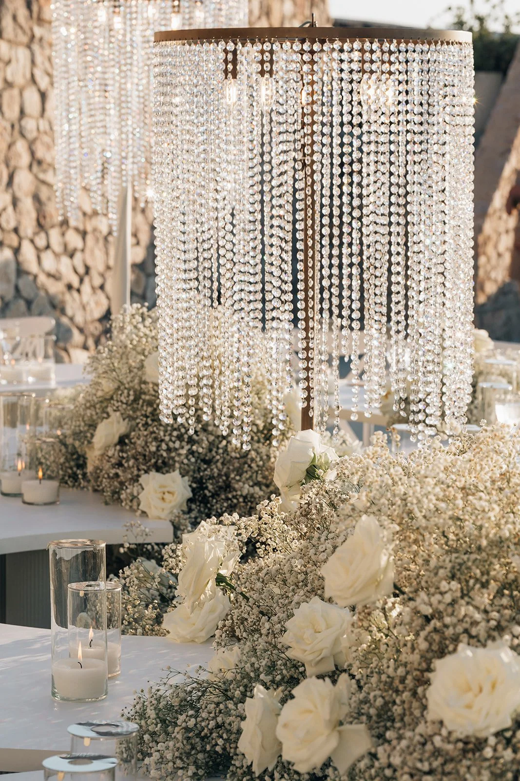 Detailed tablescape with tall glass candles, white florals, and golden accents designed for a sophisticated Santorini wedding dinner.