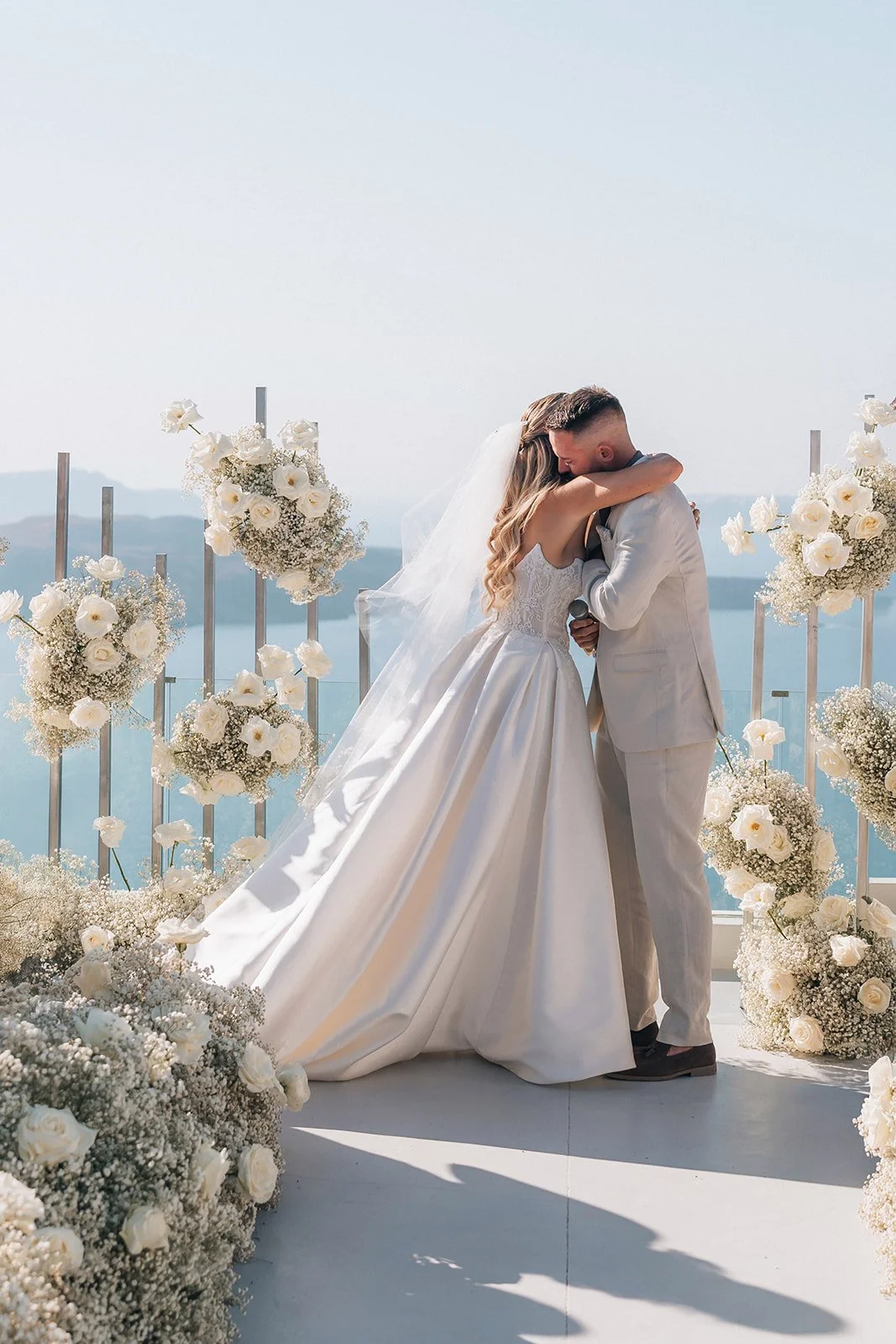 Emotional first kiss of the newlyweds during their romantic Santorini wedding ceremony surrounded by elegant white flowers.