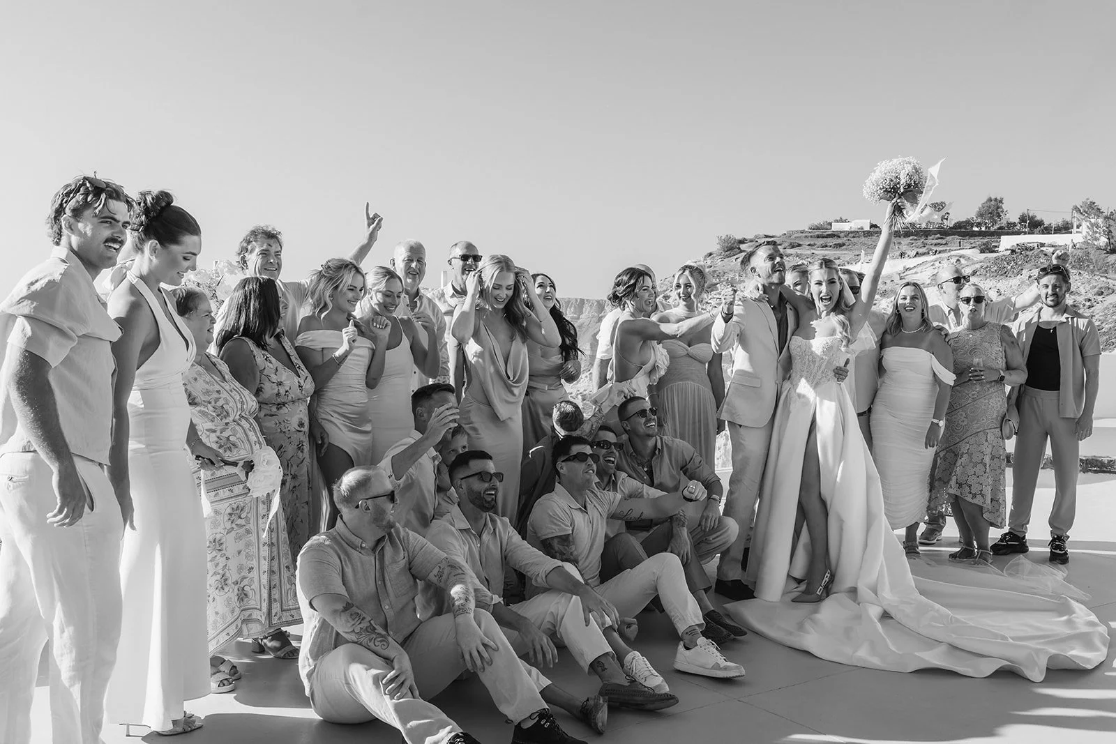 A joyful wedding group photo with guests dressed in elegant white tones, celebrating love against the breathtaking Santorini backdrop.